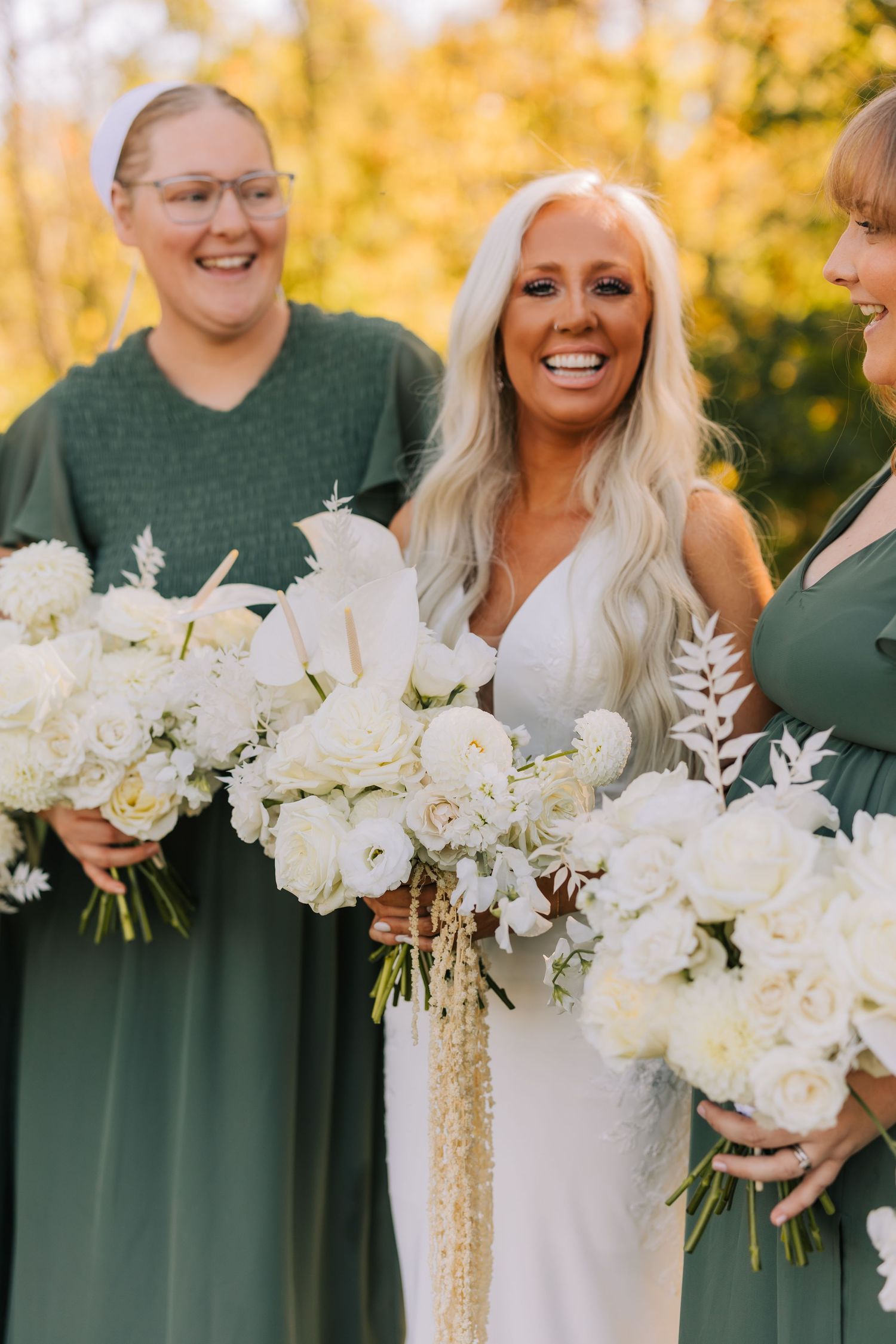 Wedding party members in green dresses hold elegant white floral bouquets during outdoor autumn photoshoot.