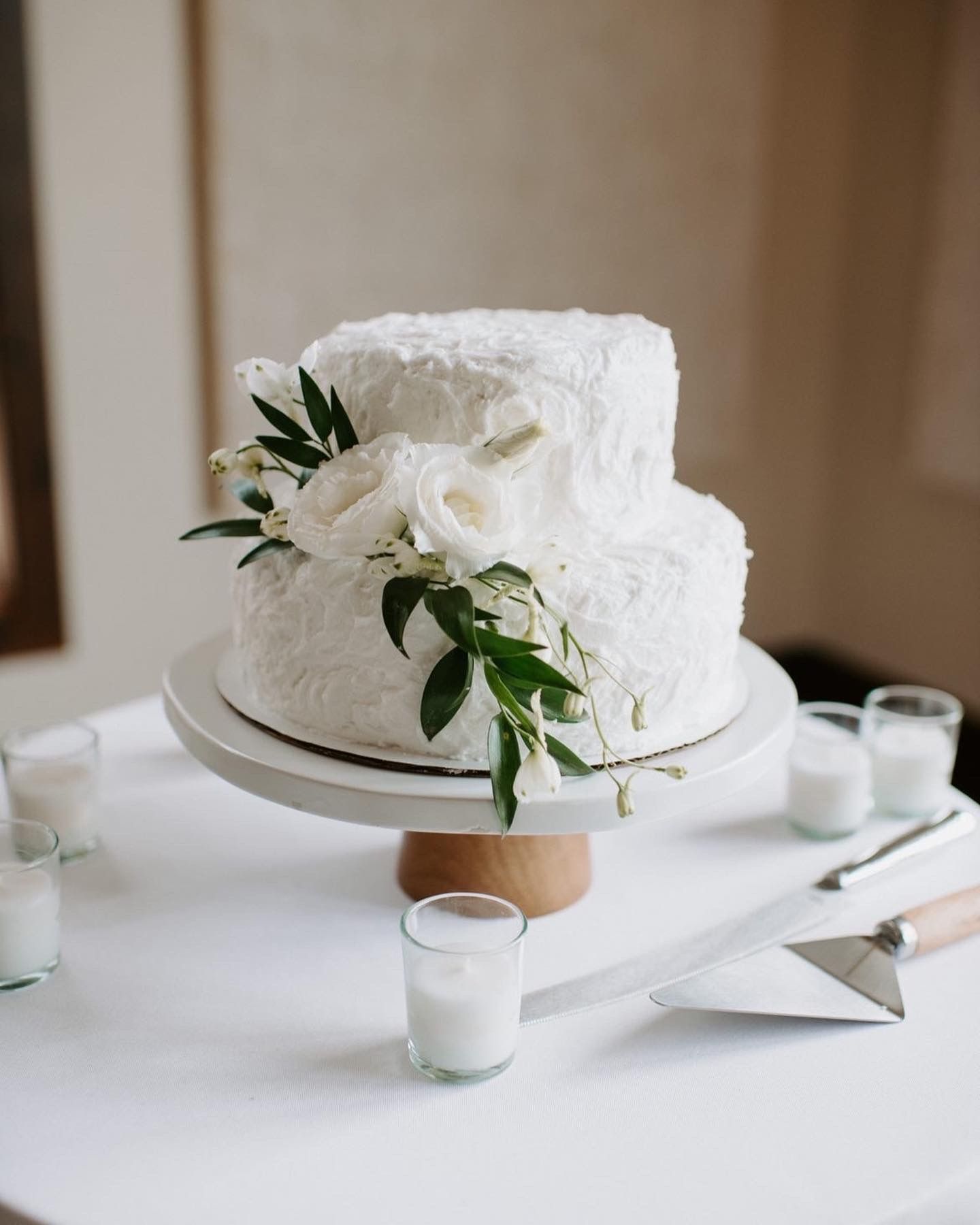 Two-tier white wedding cake decorated with fresh greenery and roses on wooden cake stand.