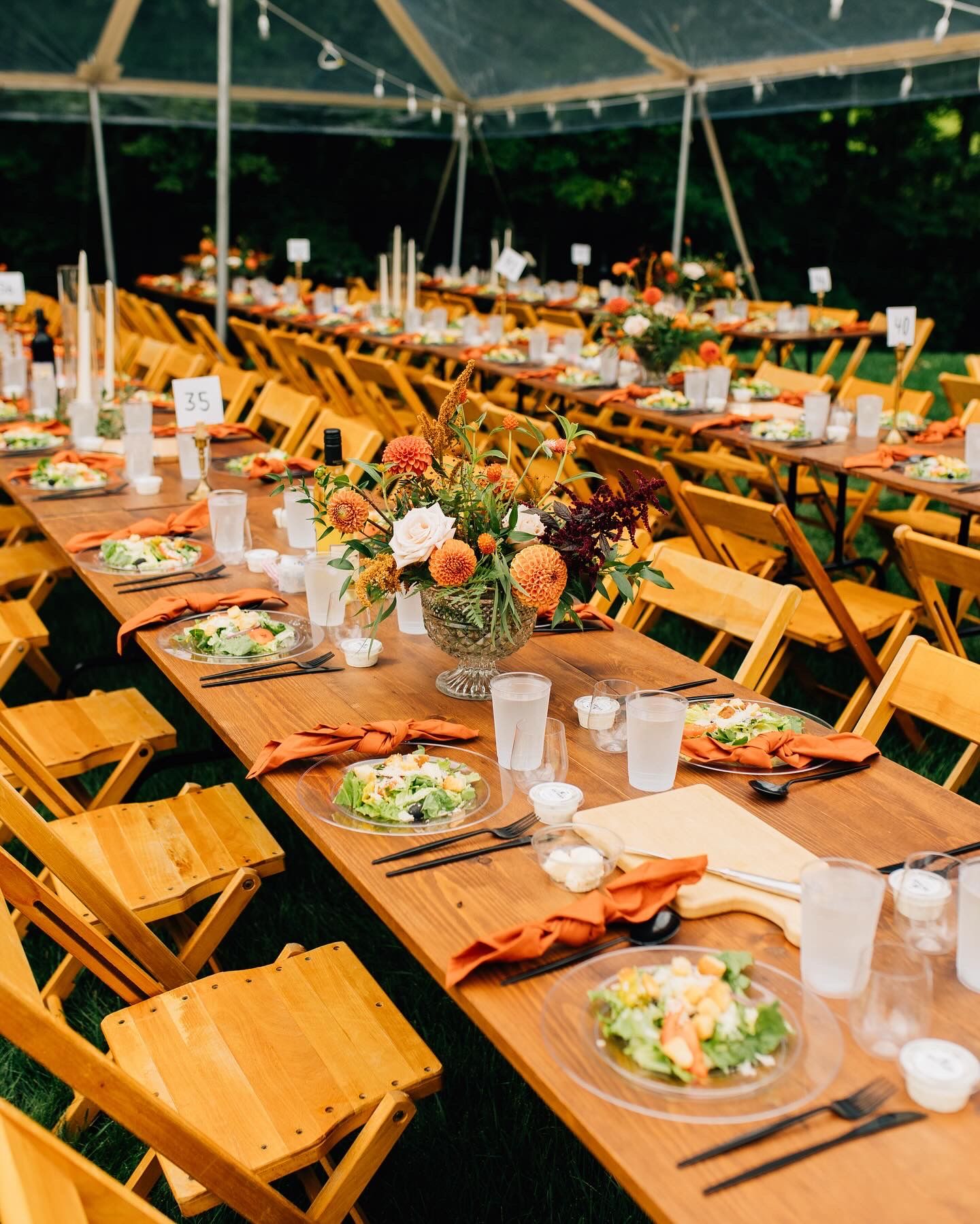 Wedding reception setup with wooden folding chairs and floral centerpieces under outdoor tent.