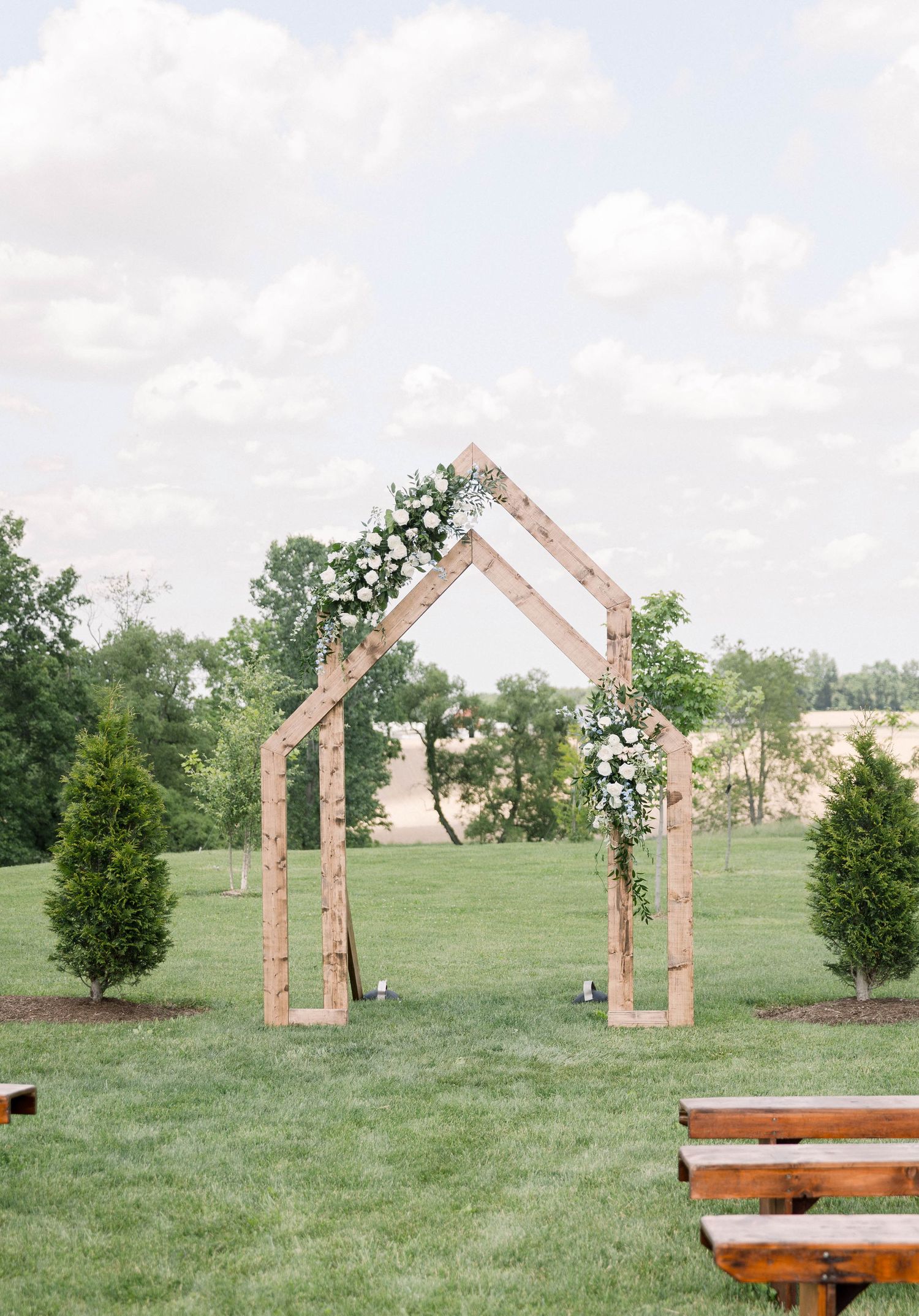 Minimalist wooden arch in chapel shape decorated with white floral arrangements in an open field.