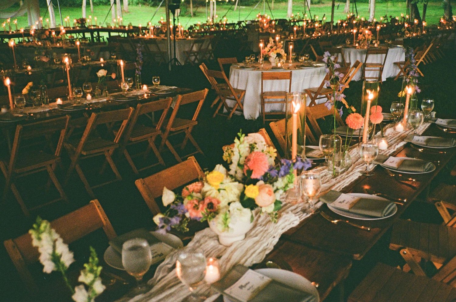 A rustic outdoor wedding reception setup with wooden tables and chairs decorated with floral centerpieces at dusk.