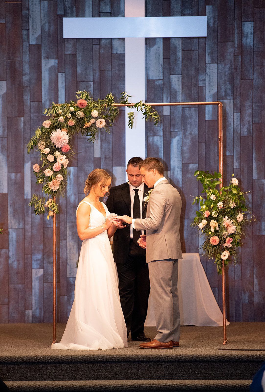 Wedding ceremony under wooden cross backdrop with copper arch and pink floral decorations.