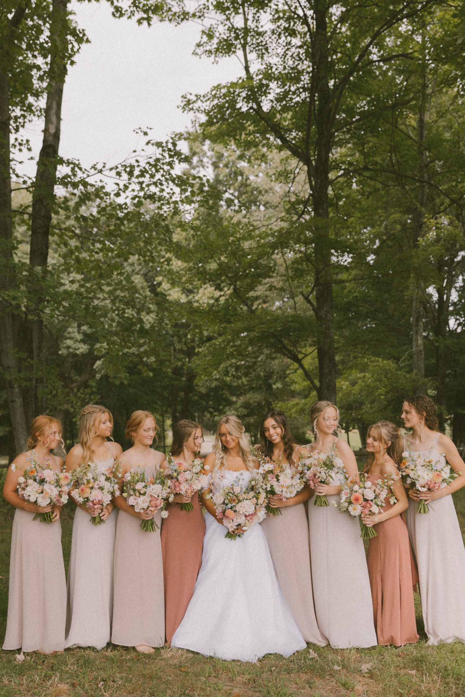 Bridesmaids in blush and mauve dresses pose with bride in a forest setting.