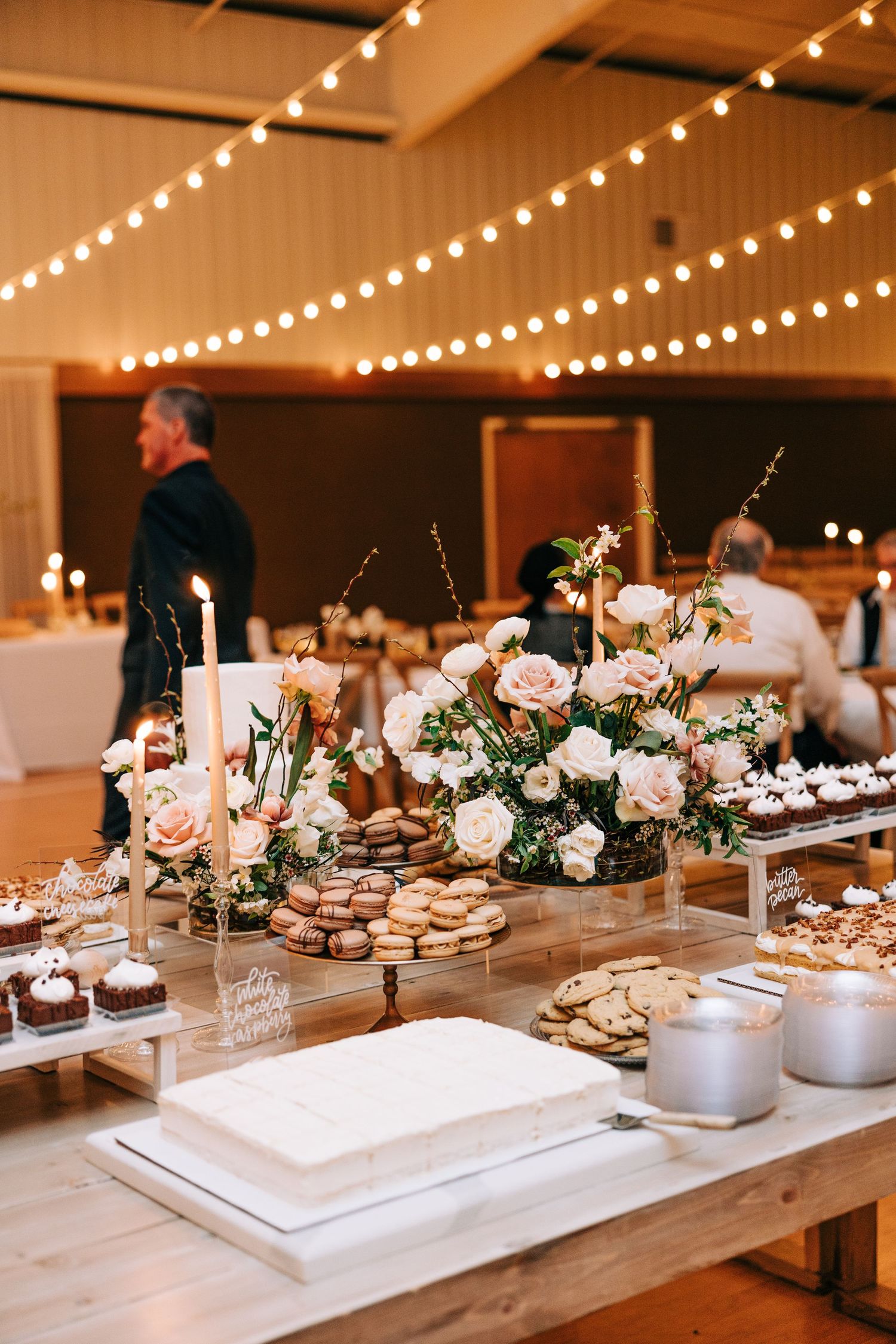 Dessert table with floral arrangements and string lights at an indoor reception venue.