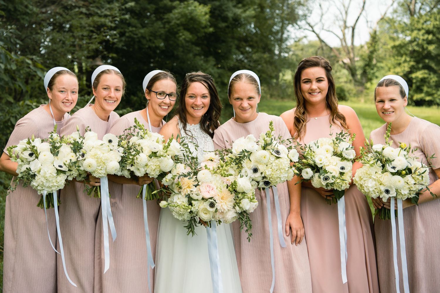 Bridesmaids in blush pink dresses hold matching white and cream floral bouquets.