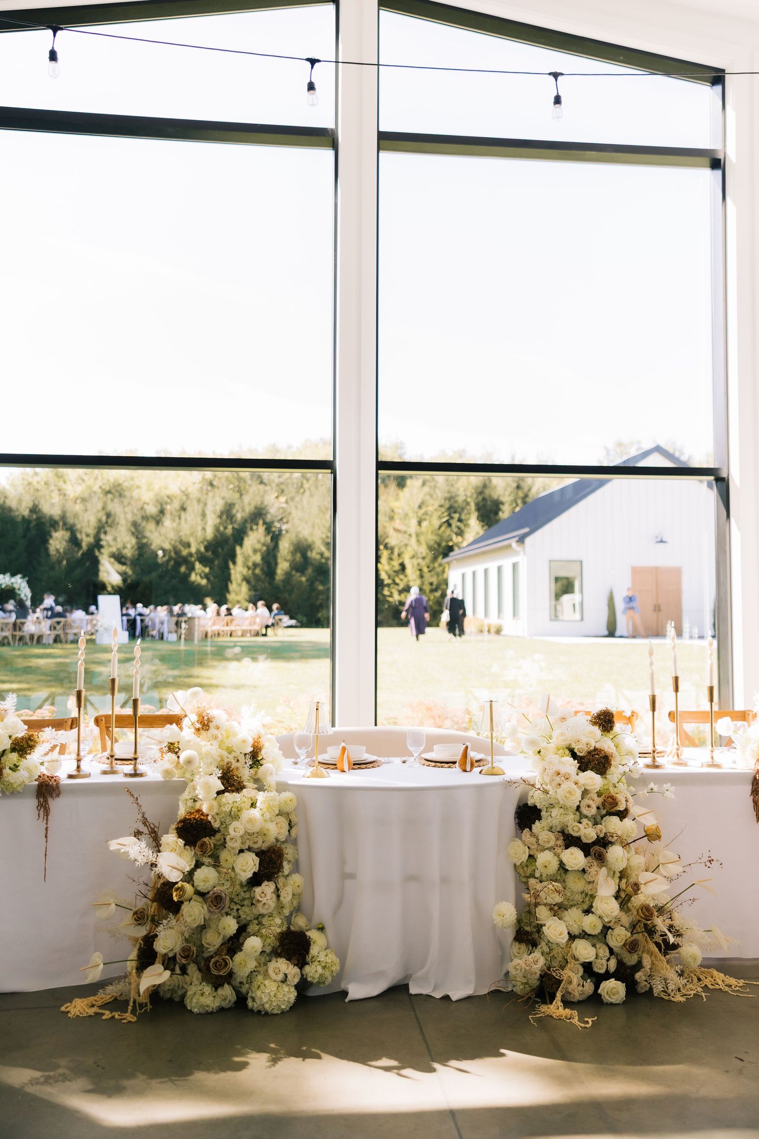 Elegant white floral arrangements with hydrangeas decorate a white-draped head table at an indoor wedding reception.