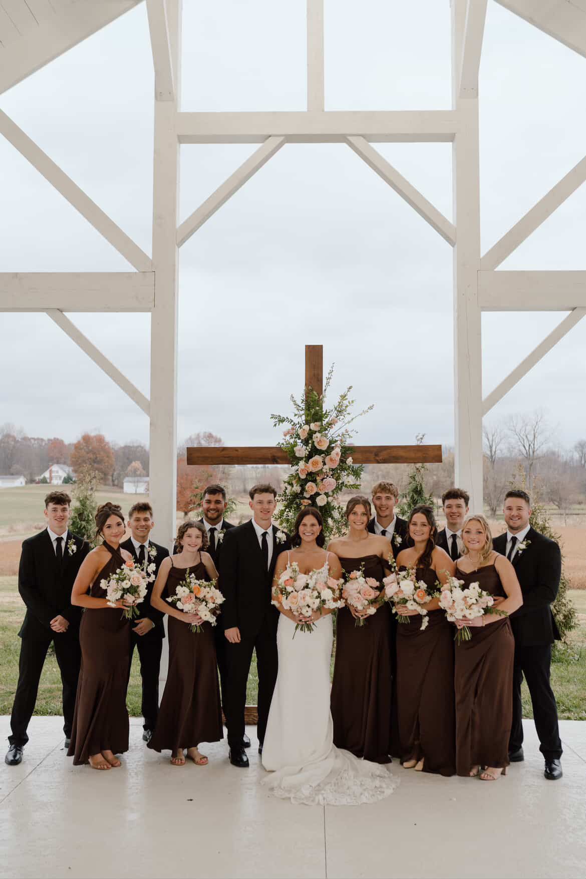 Wedding party poses together in an elegant white barn venue with rustic wooden beams and natural light.