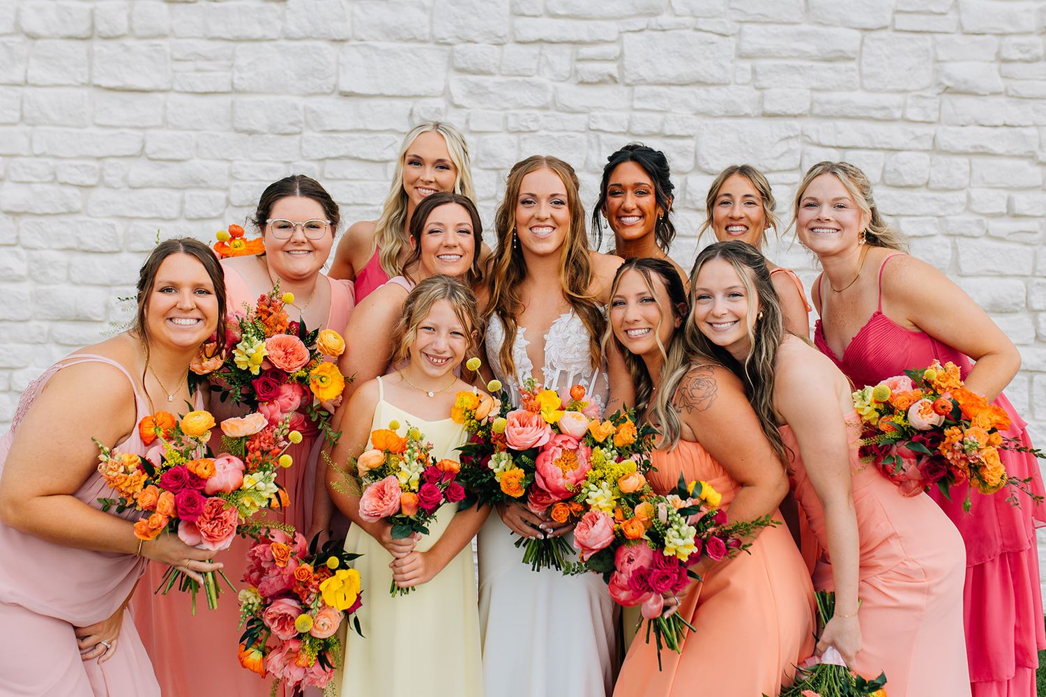 Wedding party in coral and pink dresses holding vibrant orange and pink floral bouquets against white brick wall.