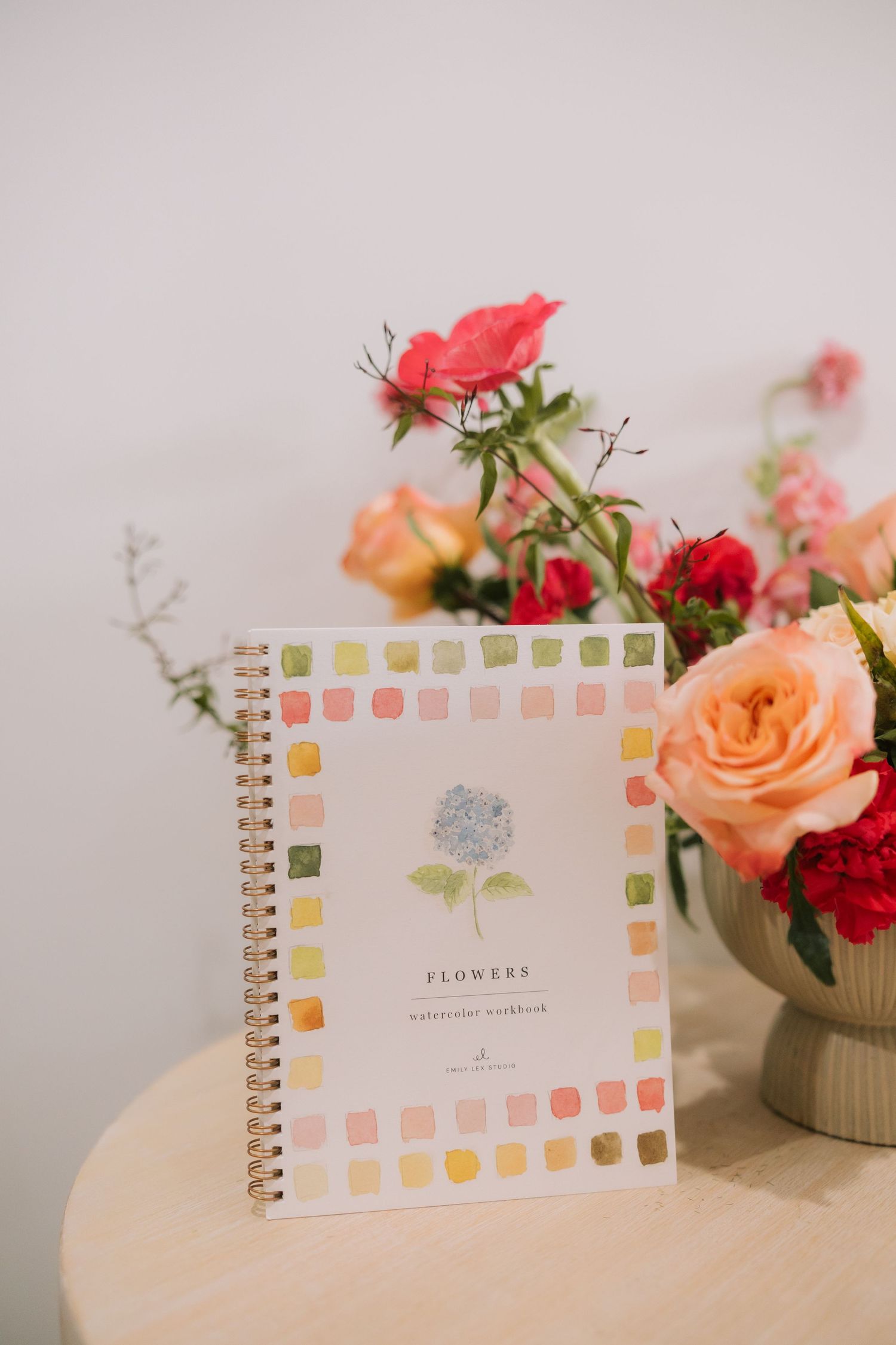 A spiral notebook with floral design sits beside fresh roses and carnations in peach and red tones on a light surface.