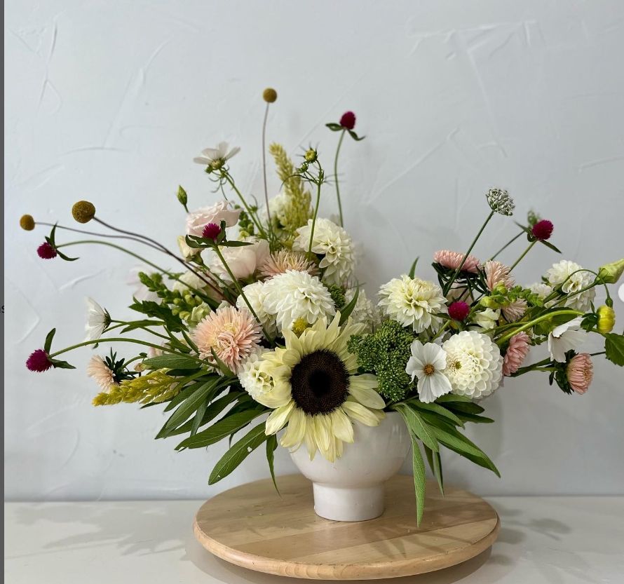 Whimsical arrangement with sunflowers, white dahlias, and unique stems in a white ceramic vessel.