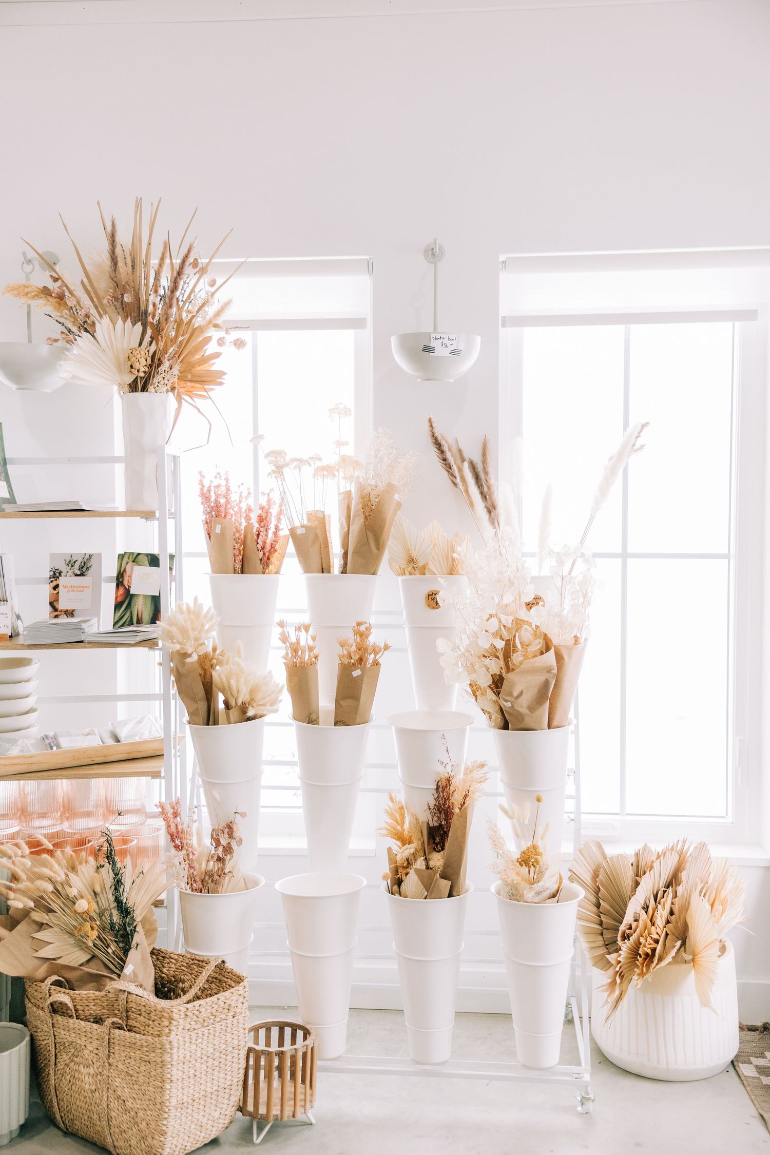 Dried flowers and pampas grass displayed in white vases on shelving in a bright sunlit room.