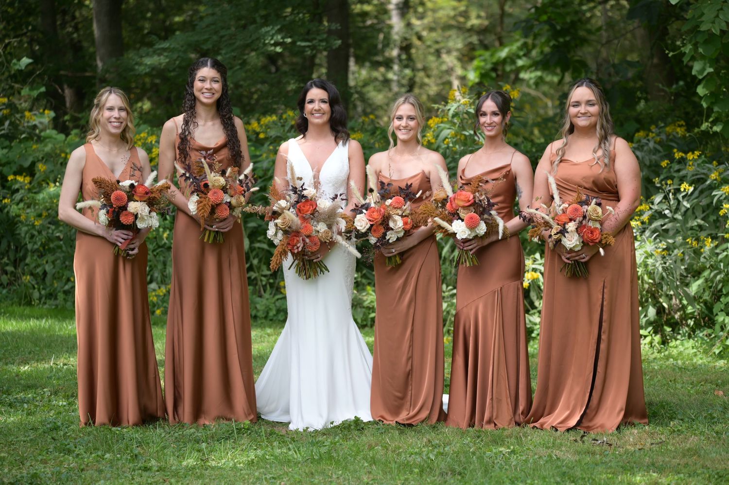 Wedding party in rust-colored dresses poses together in a garden setting with lush greenery and flowers.