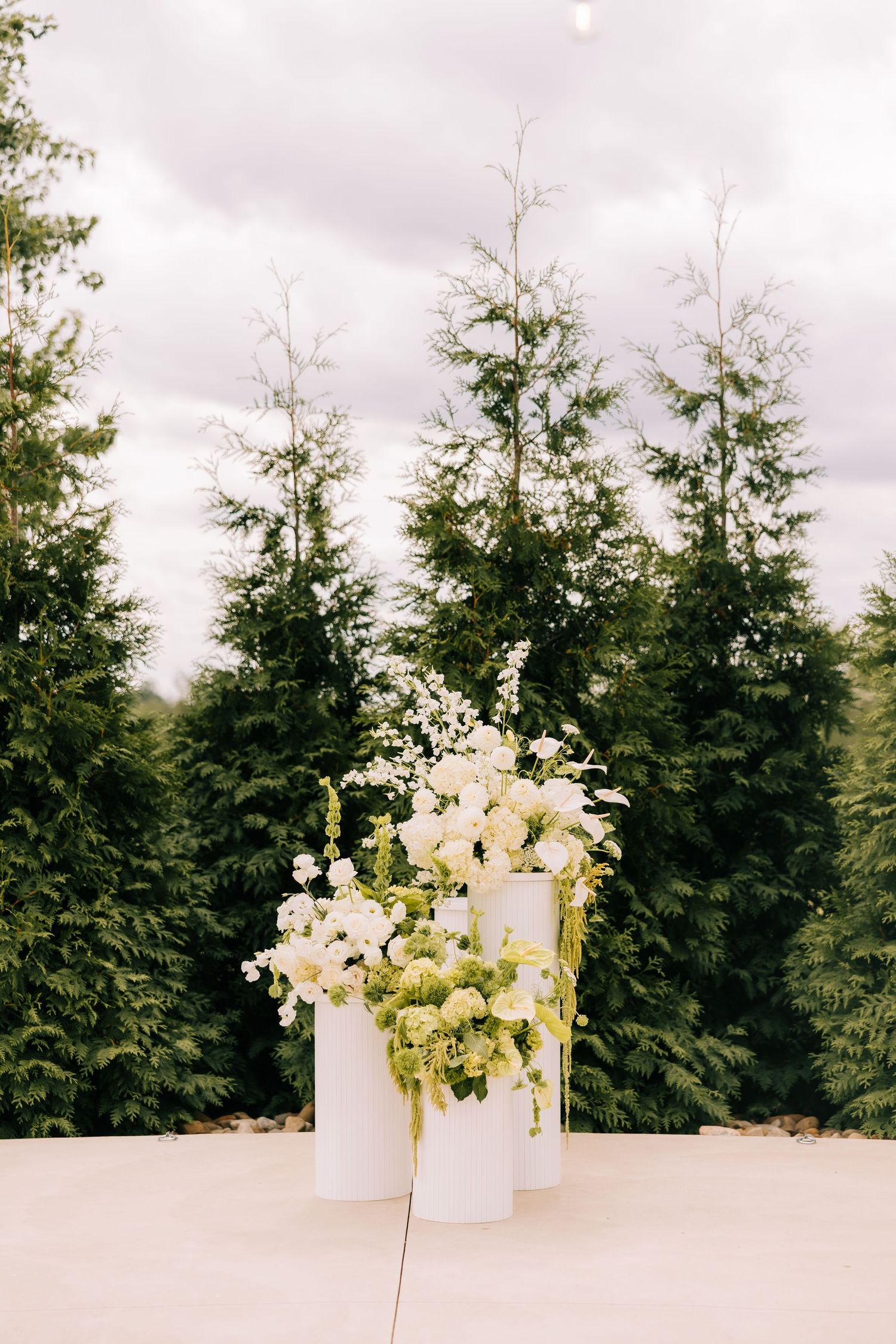 White floral arrangements in tall vases against a backdrop of evergreen trees under cloudy skies.