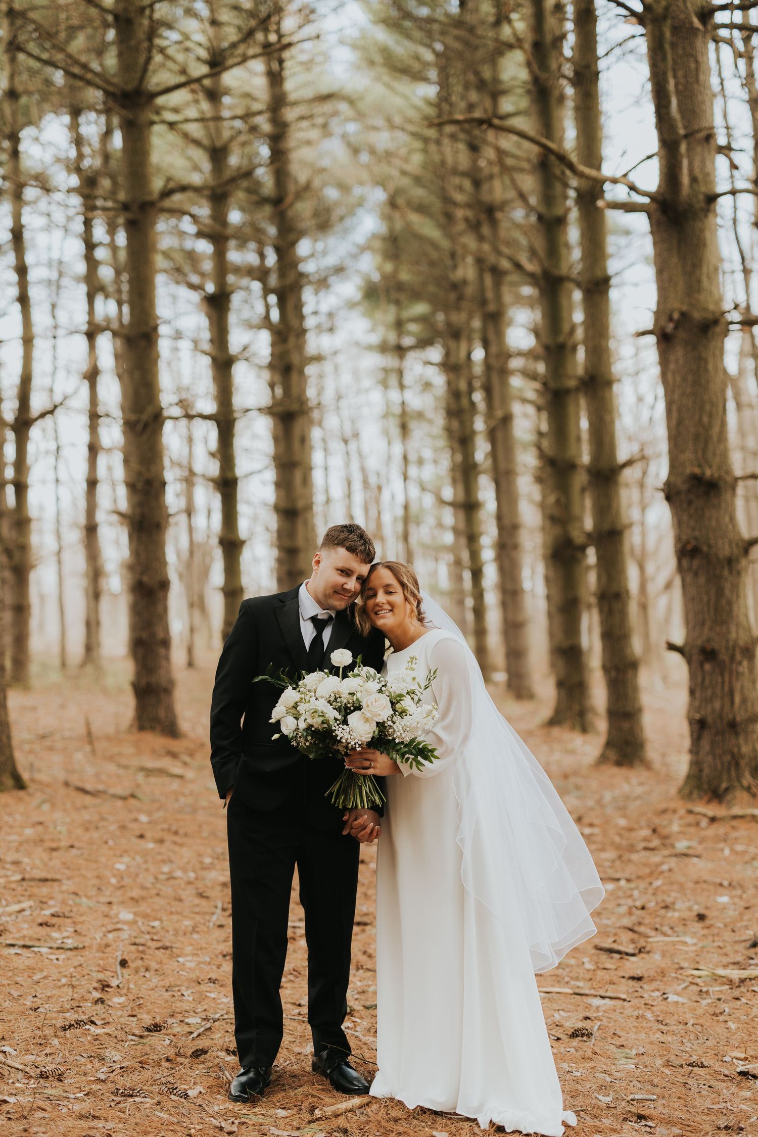 A romantic wedding couple shares an intimate moment among tall pine trees on a forest floor covered in fallen leaves.