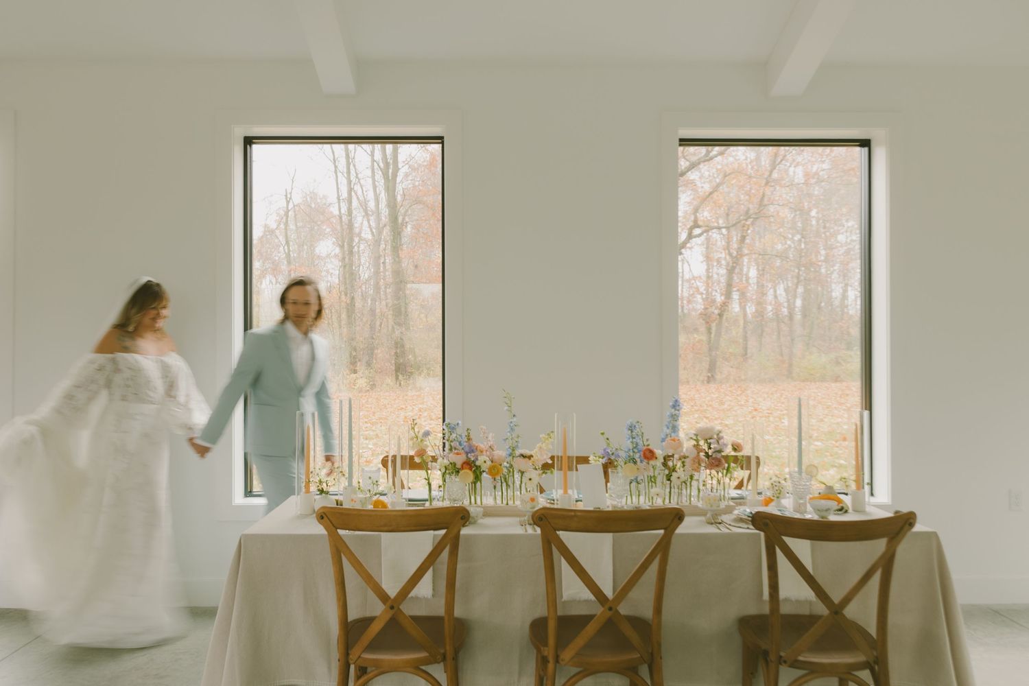 Minimalist wedding reception setup with cross-back chairs and floral table decorations near large windows.