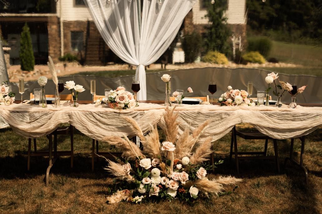 Elegant outdoor wedding table setting with white draping and neutral floral arrangements on a rustic tablescape.