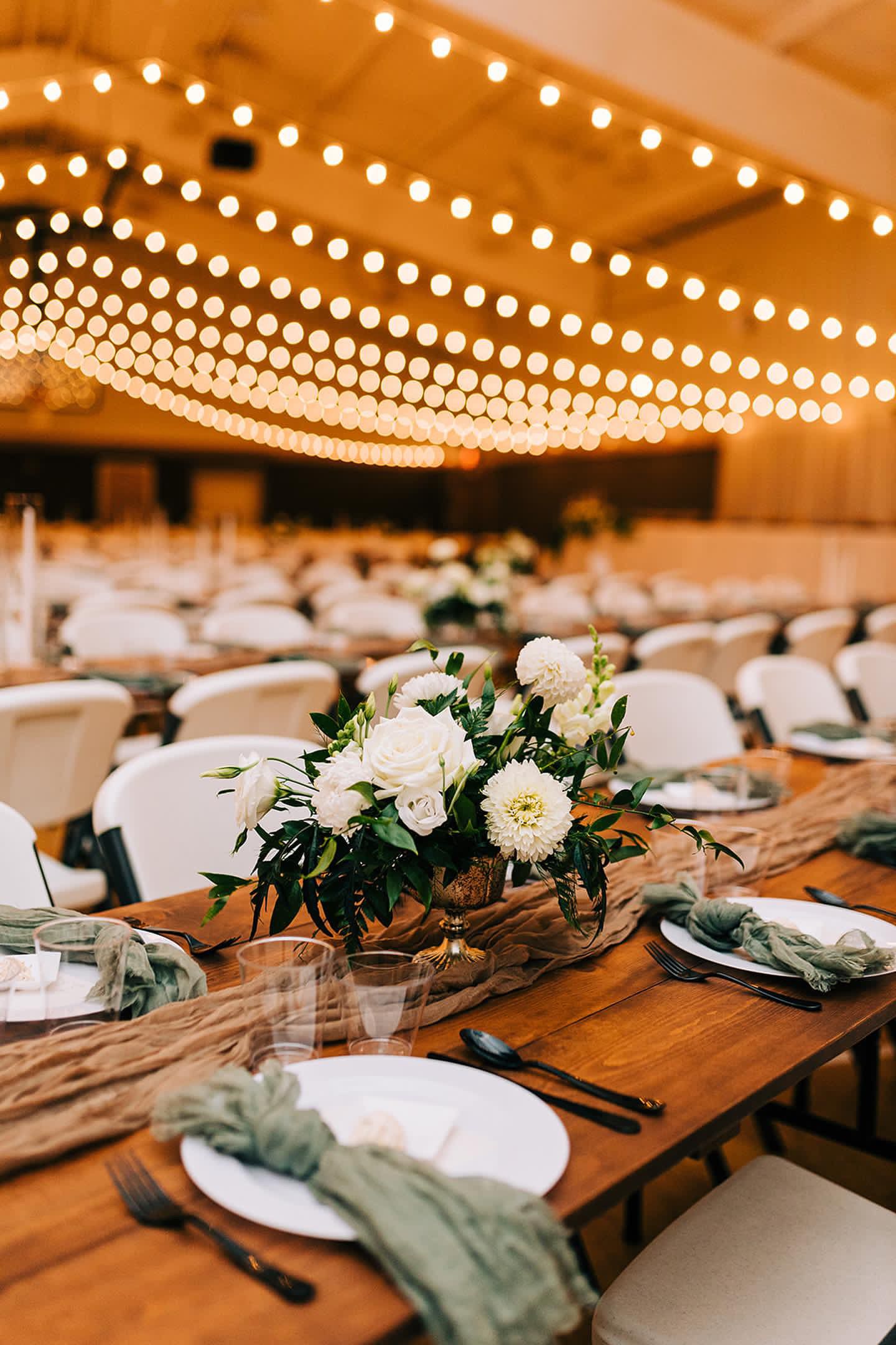 Reception table setting with white floral centerpiece under warm string light canopy.