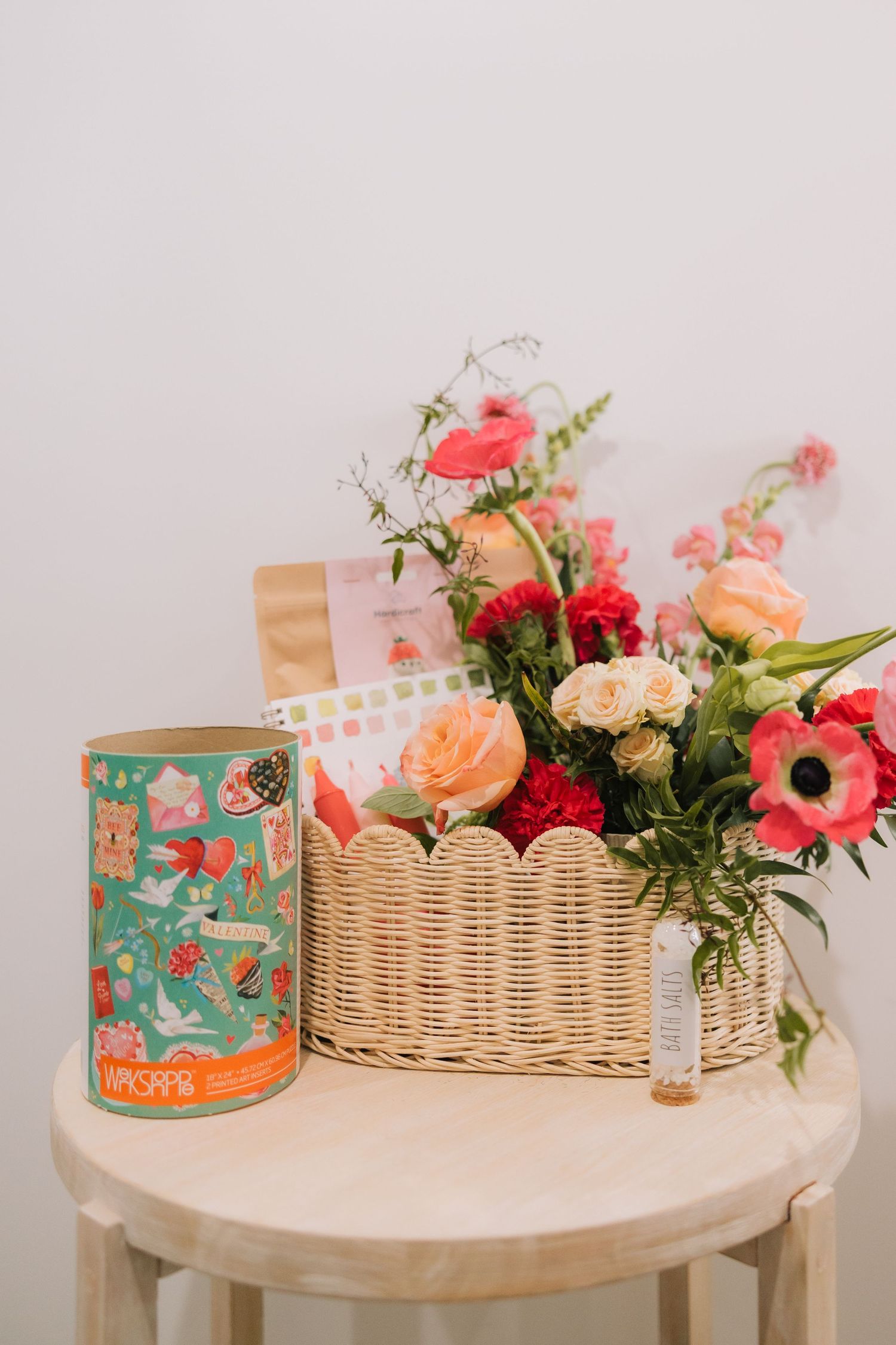 A round wooden table displays a wicker basket filled with pink and red flowers alongside decorative floral-patterned tins.