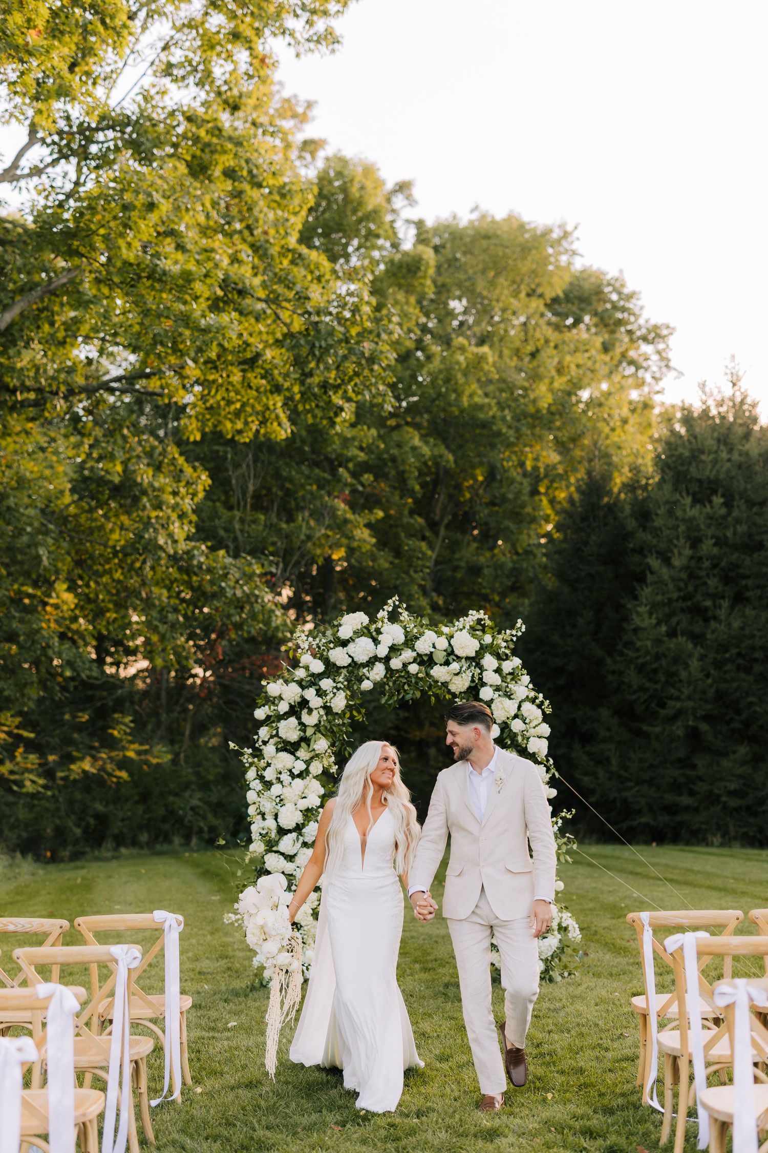A romantic outdoor wedding ceremony setup with white floral arch and wooden chairs on a lush green lawn at sunset.