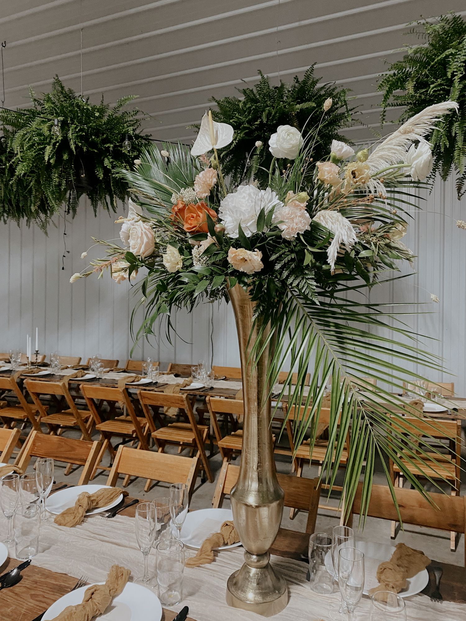 Tall white and peach floral centerpiece with palm fronds in gold vase surrounded by wooden chairs.