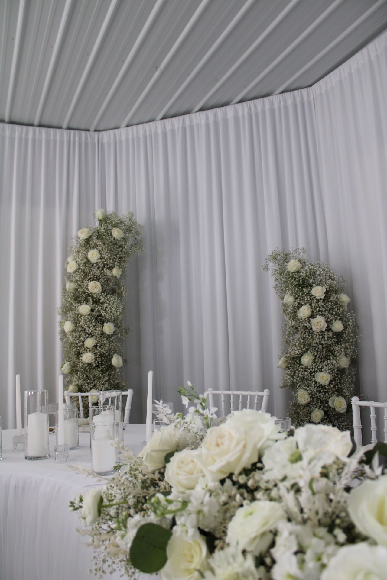 All-white wedding reception decor with tall floral arrangements against draped fabric backdrop.