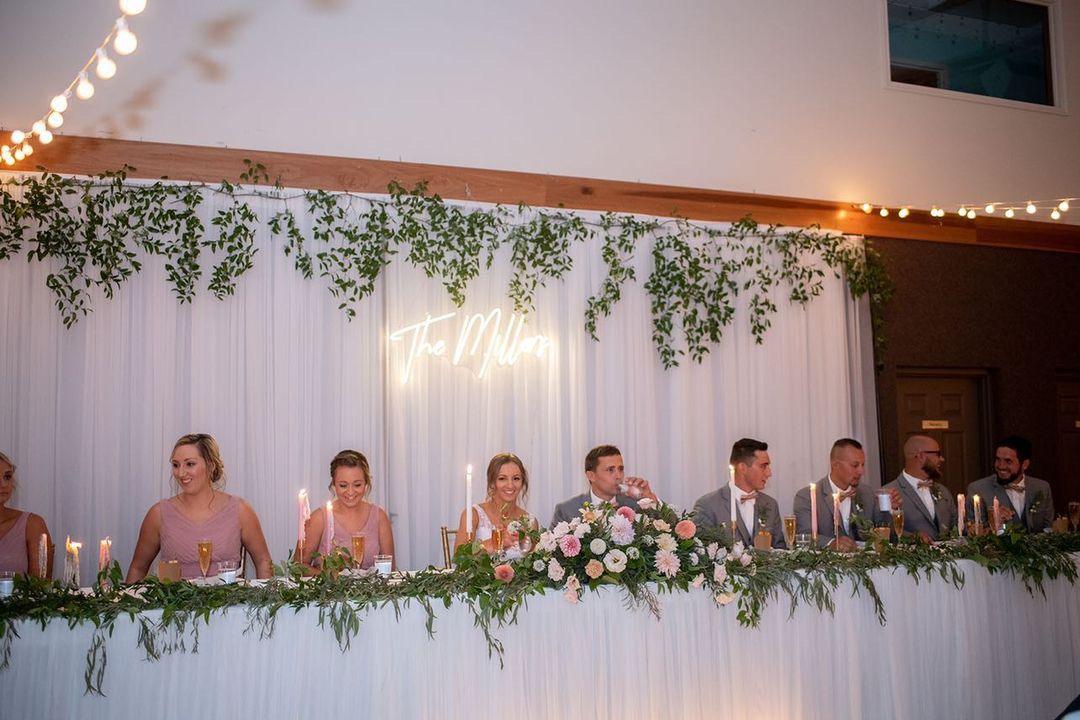 Wedding party seated at elegant head table decorated with greenery and a neon sign against white backdrop.