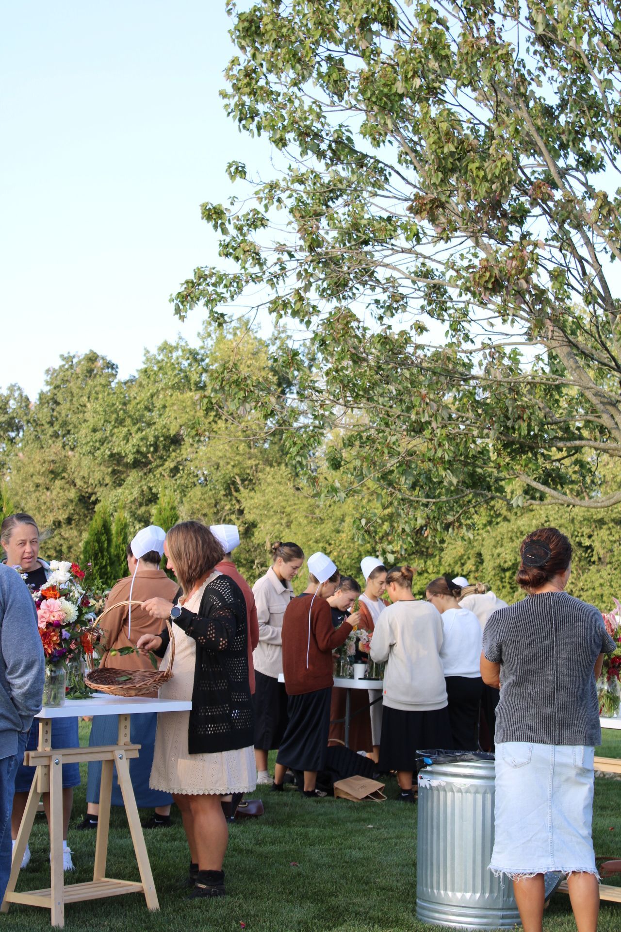 Outdoor gathering with people standing near tables under trees.