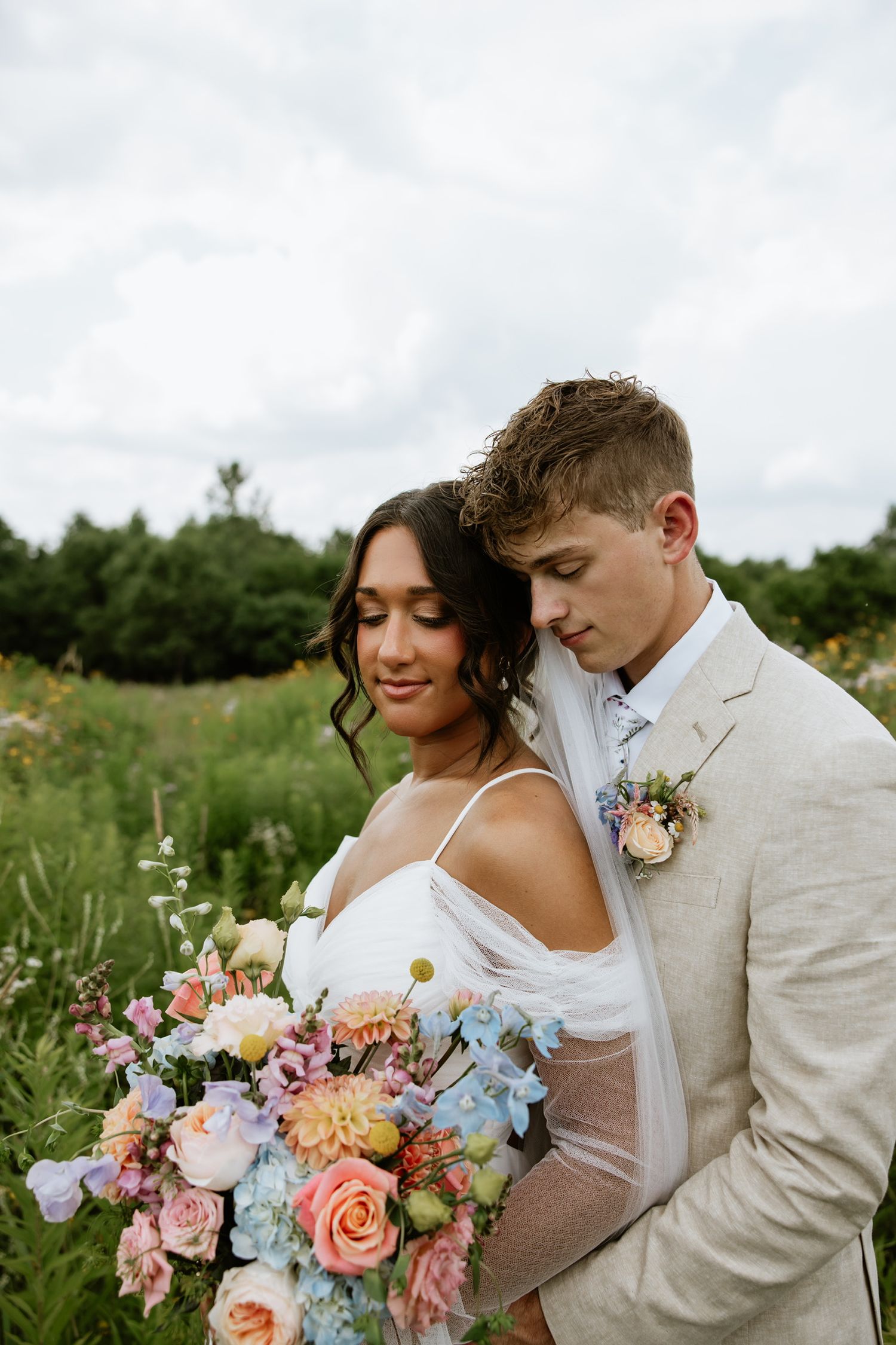 Couple in formal attire poses with stunning pastel floral bouquet in wildflower meadow.