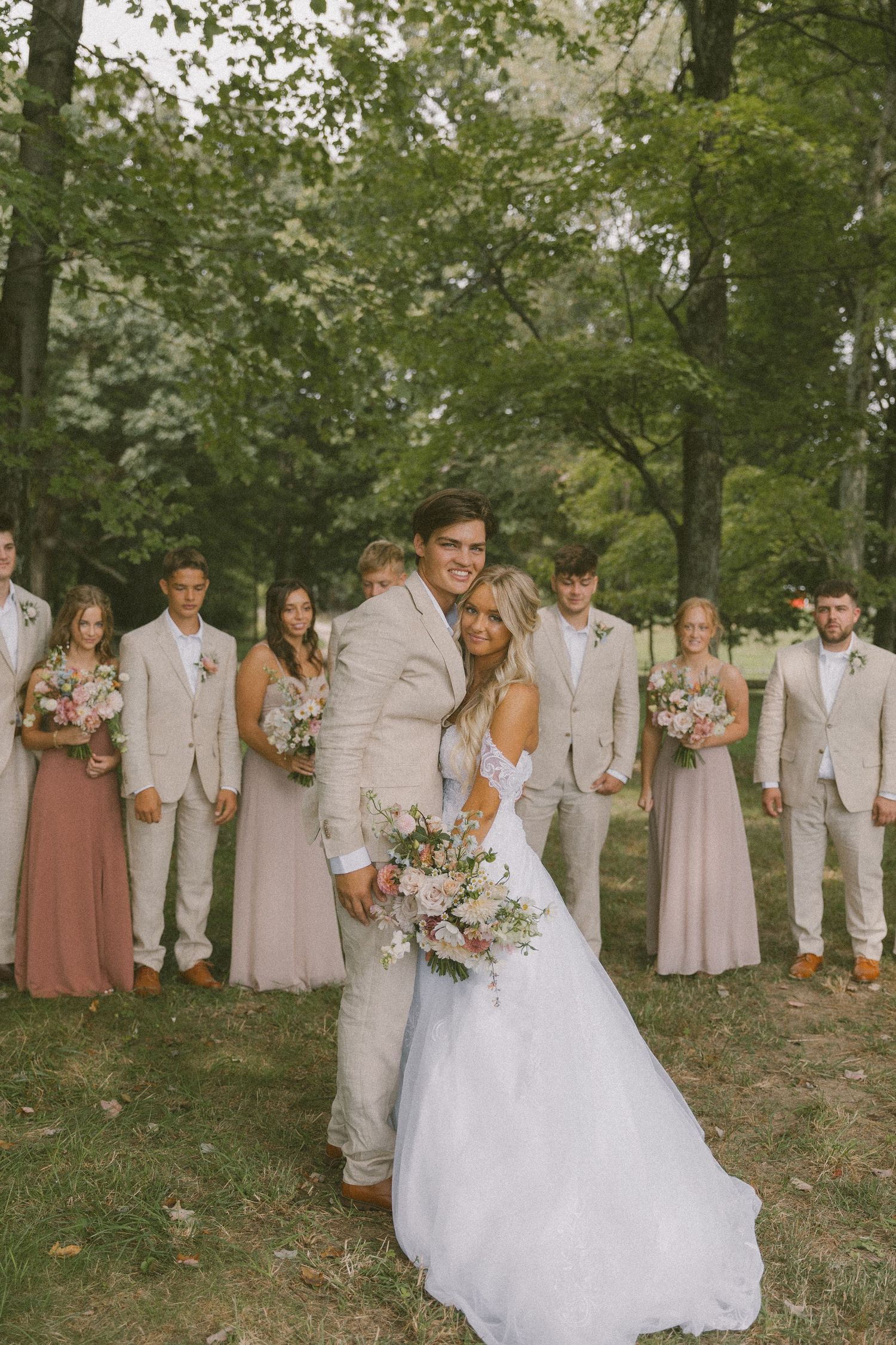 Wedding party in light neutral attire poses together in a wooded outdoor setting.