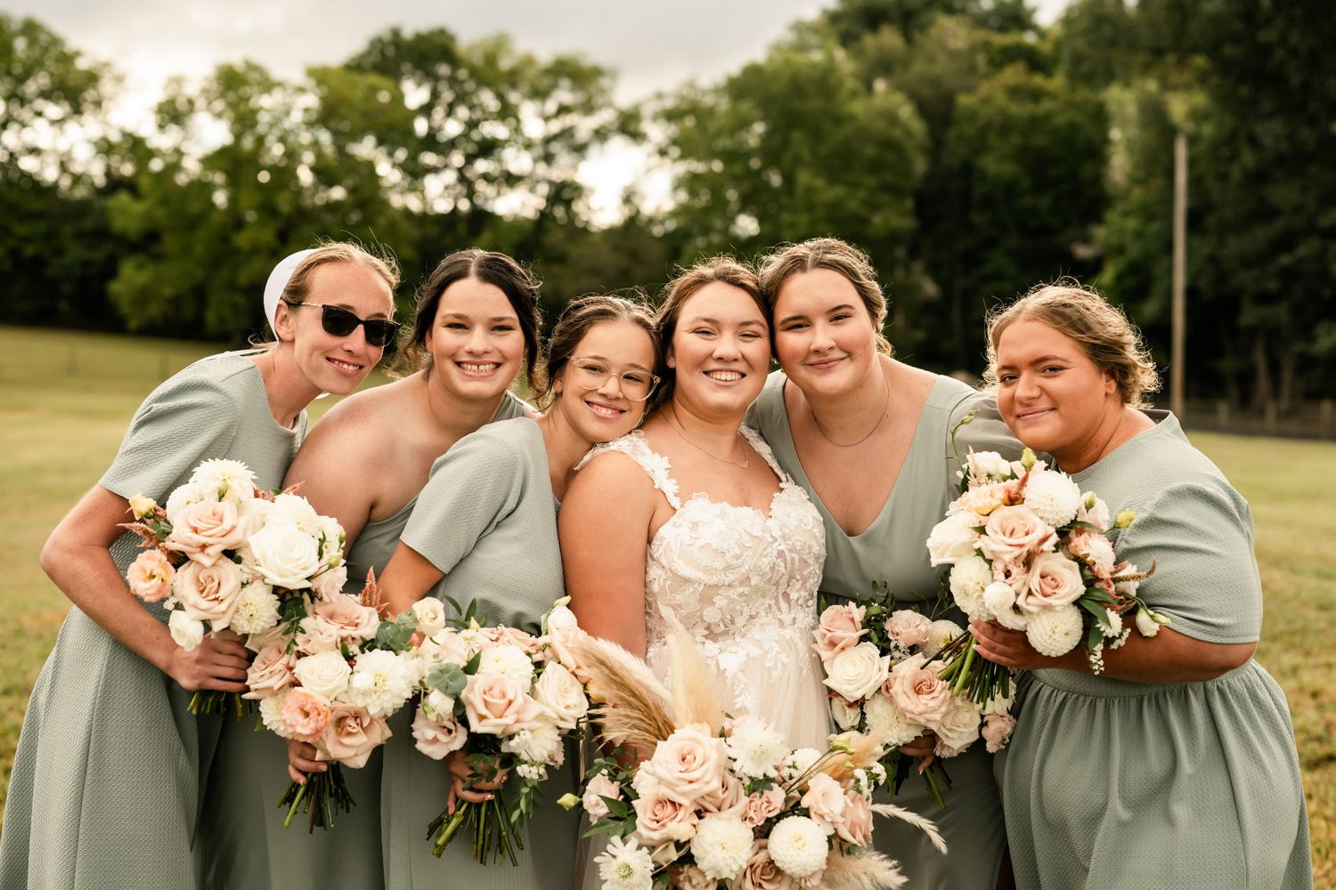 Joyful wedding party in sage green dresses holding blush bouquets outdoors.