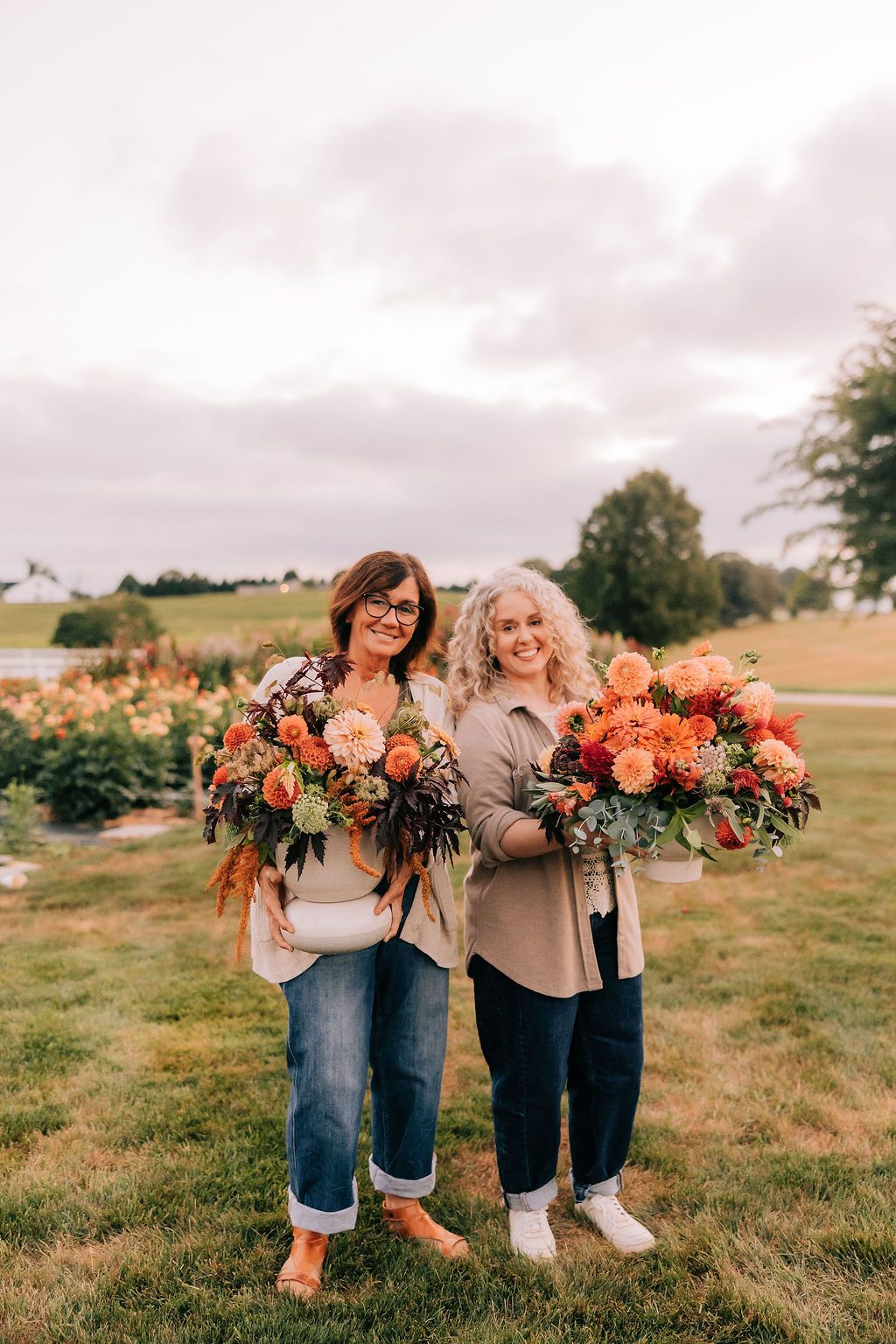 Two people holding vibrant orange and pink floral bouquets pose playfully in a grassy field at sunset.