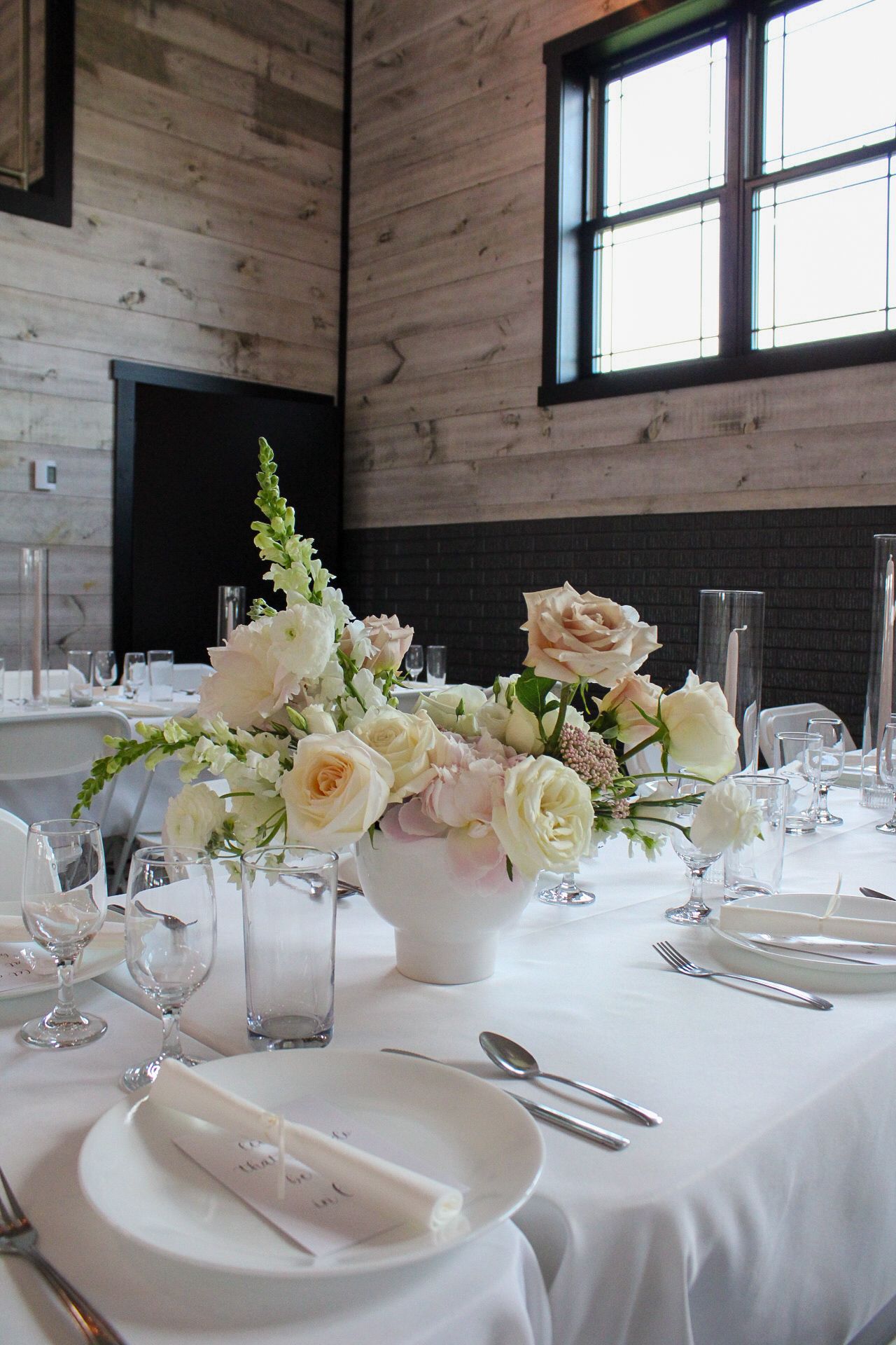 Elegant wedding reception table with white linens, blush roses, and crystal glassware in a rustic wooden venue.