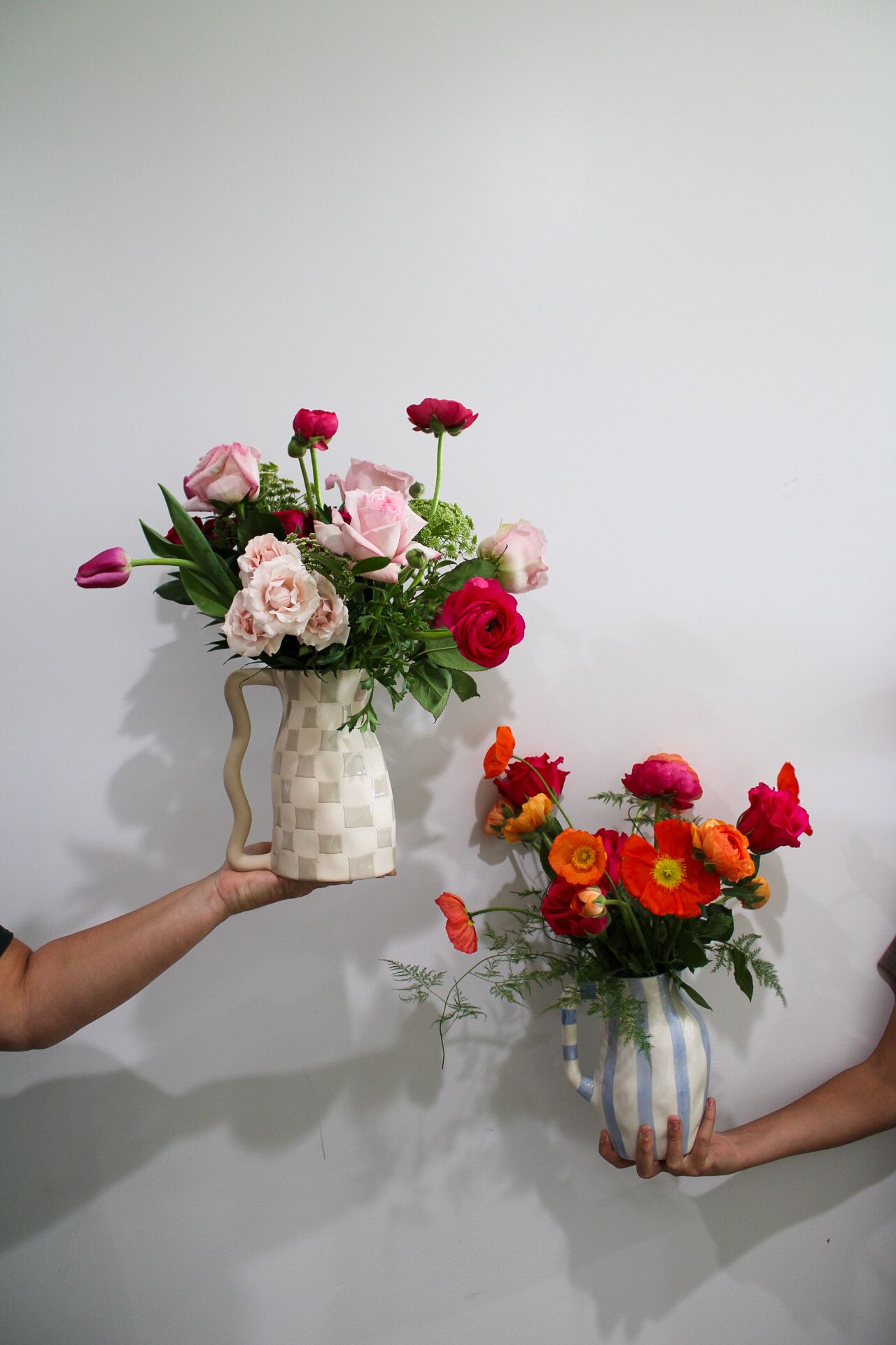 Two hands holding colorful floral arrangements in decorative vases against a white wall.