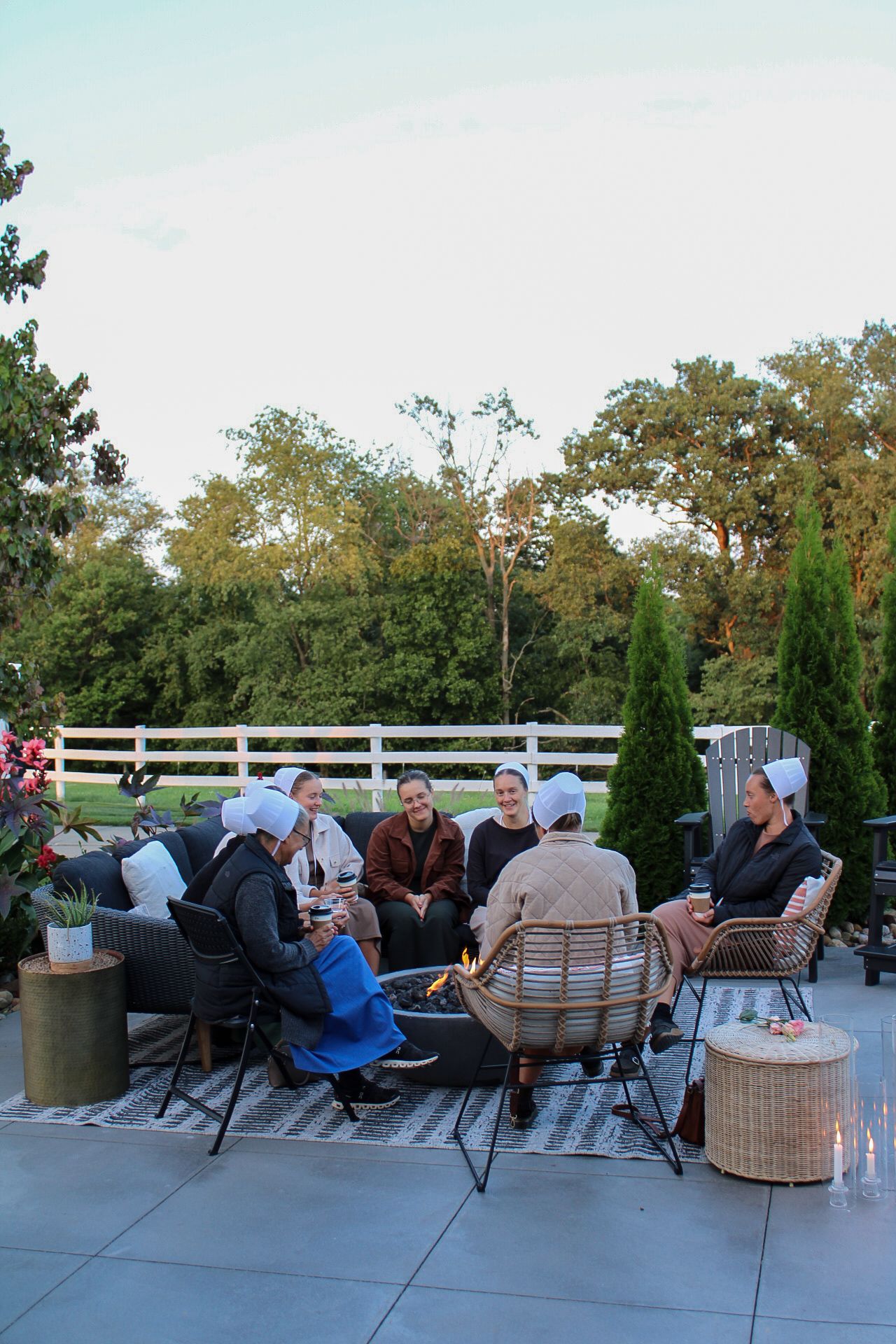 People seated in casual outdoor gathering space with landscaped garden and white fence in background.