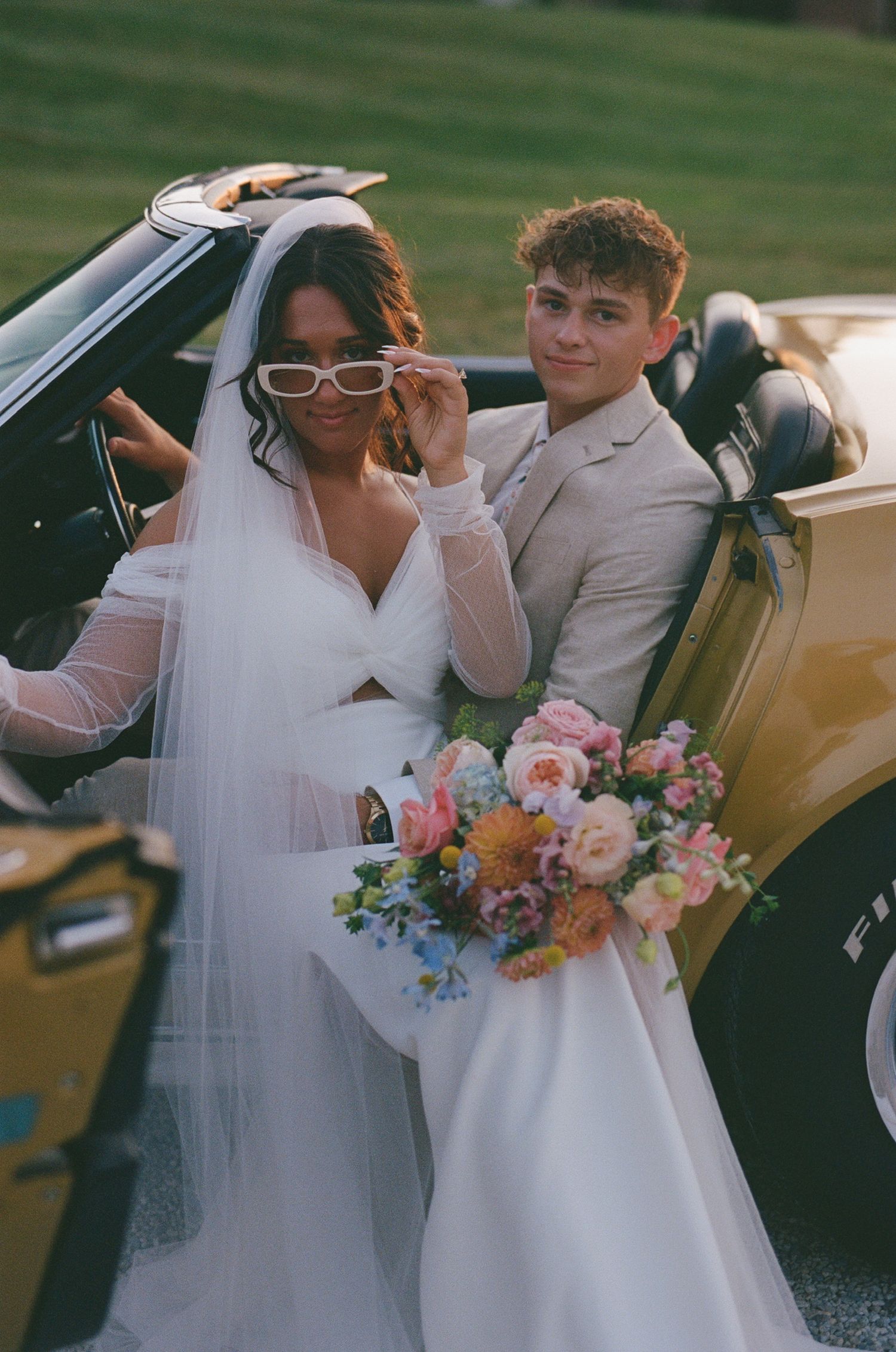 A wedding couple pose together in a vintage yellow car with a colorful bouquet of flowers.