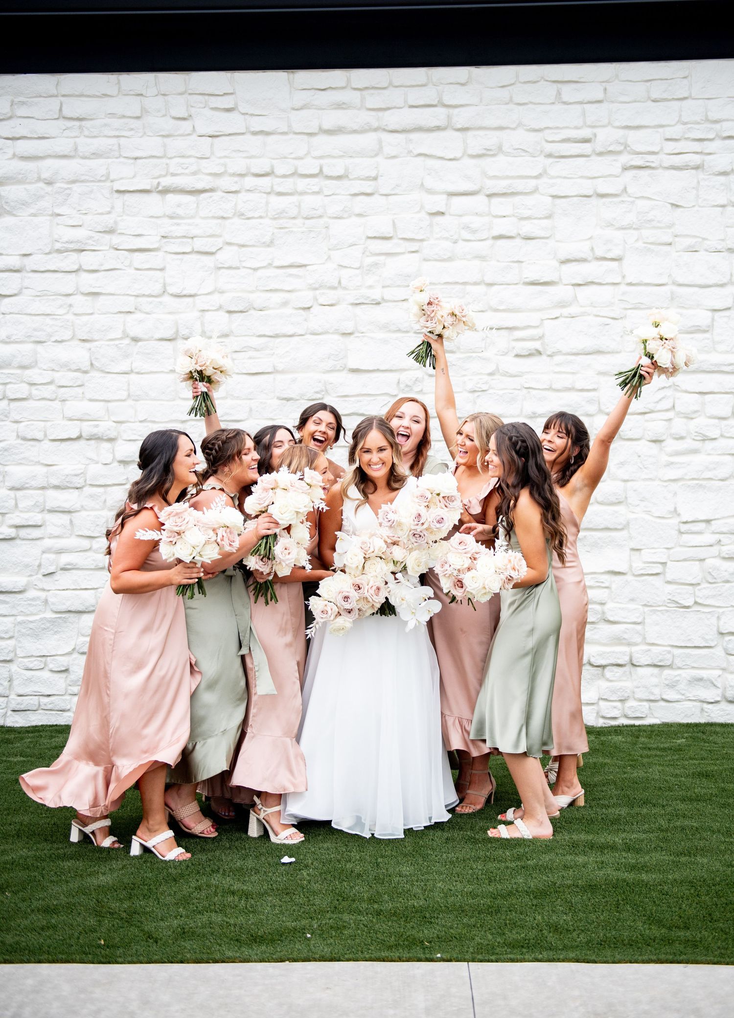 Joyful bridal party in pink and sage dresses celebrates against white brick wall.