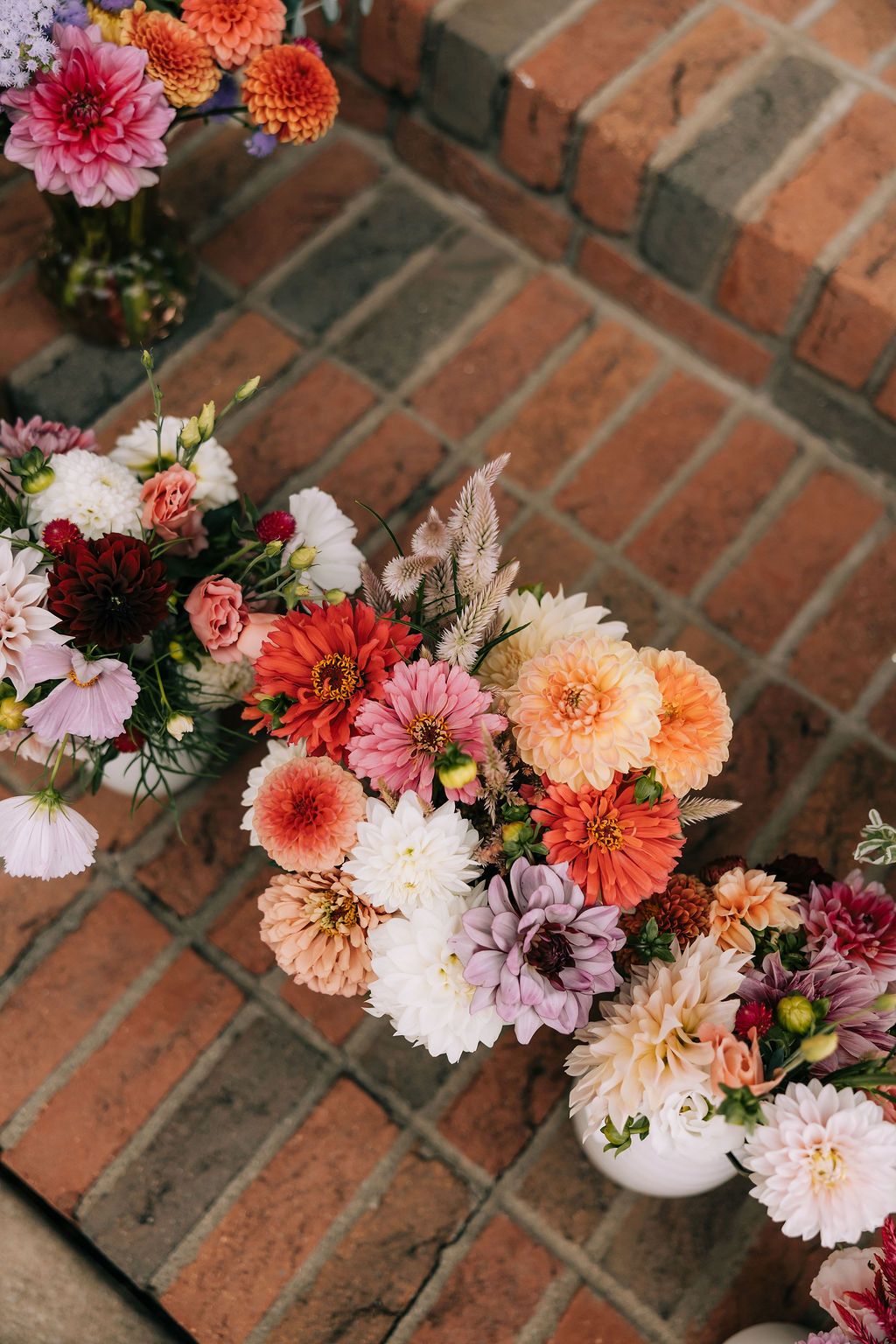 Colorful dahlias and mixed flowers arranged in white vases on a rustic brick surface create a romantic garden display.