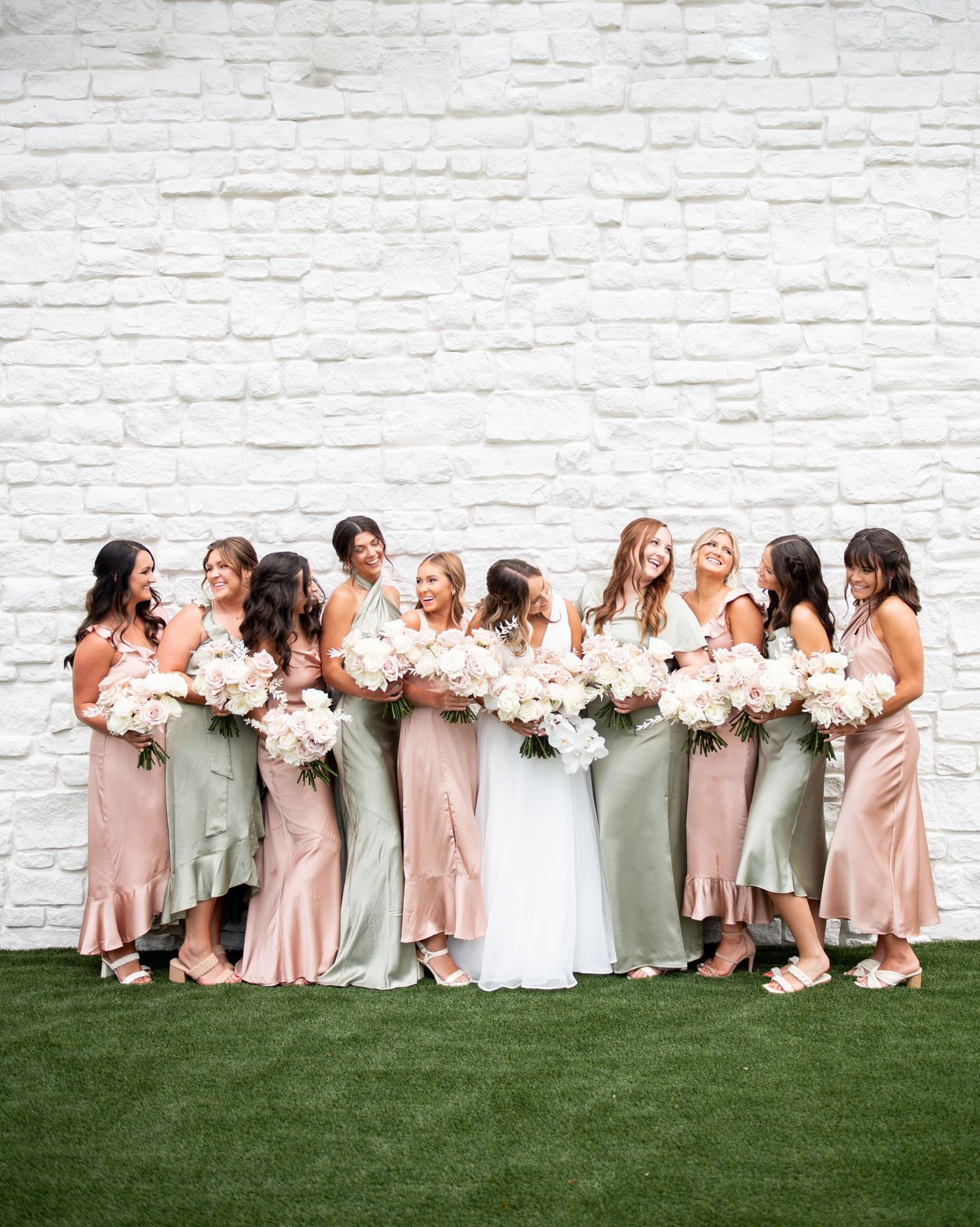 Bridal party in pastel silk dresses poses against white brick wall holding bouquets.