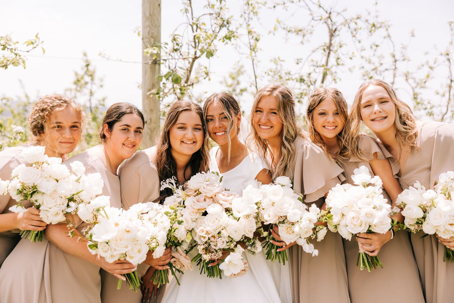 Bridal party in neutral dresses holding lush white floral bouquets outdoors.