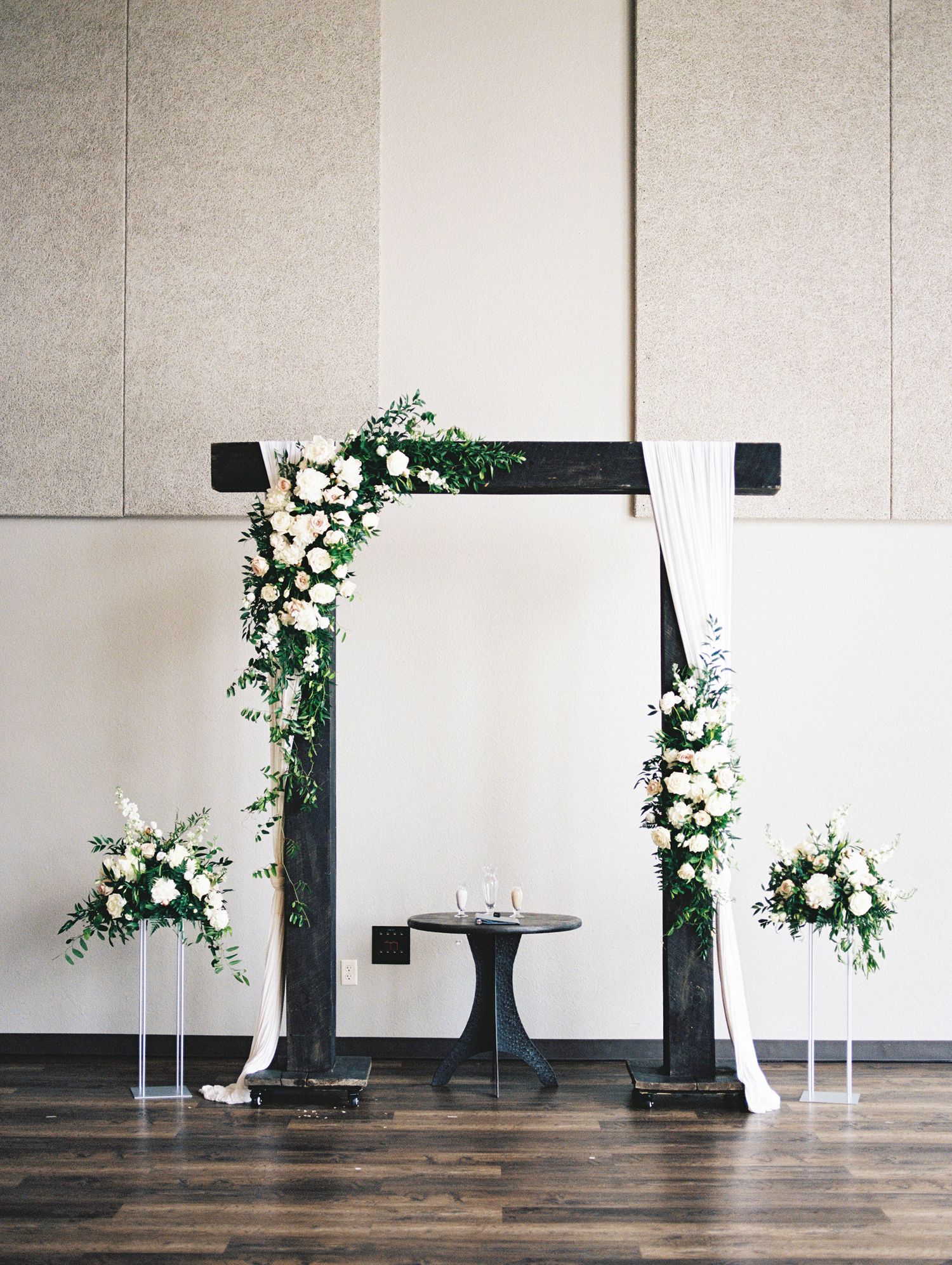 Modern black wedding arch decorated with white floral arrangements and greenery against a neutral wall.