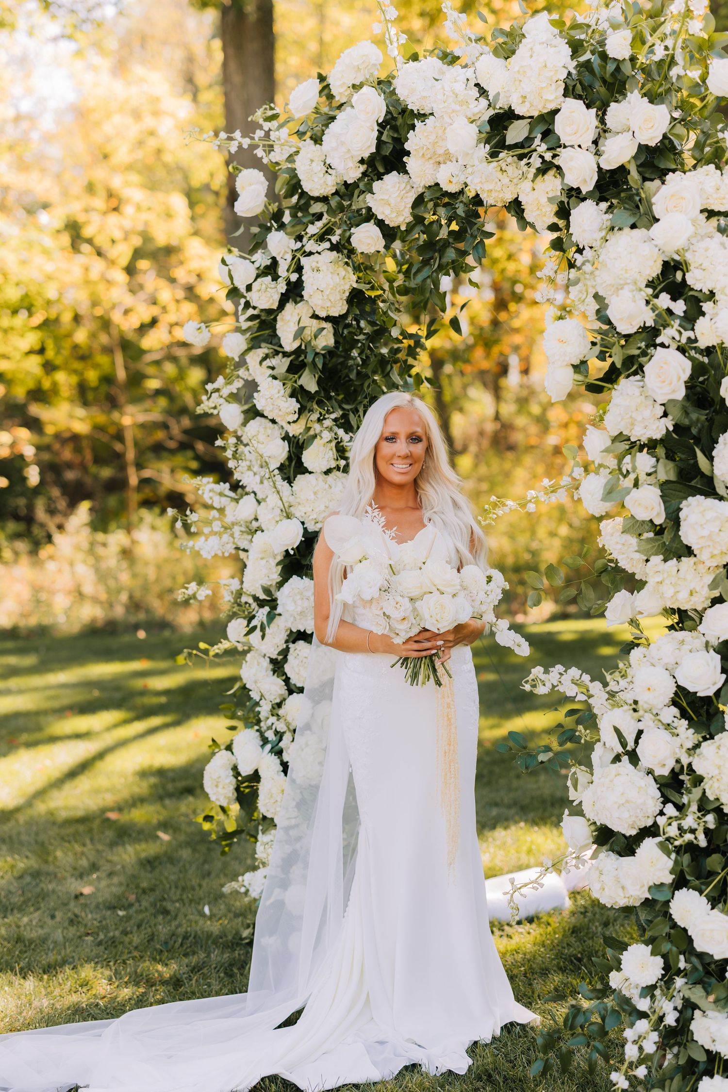 Beautiful floral wedding arch with white roses and greenery creates an elegant outdoor ceremony backdrop in golden sunlight.