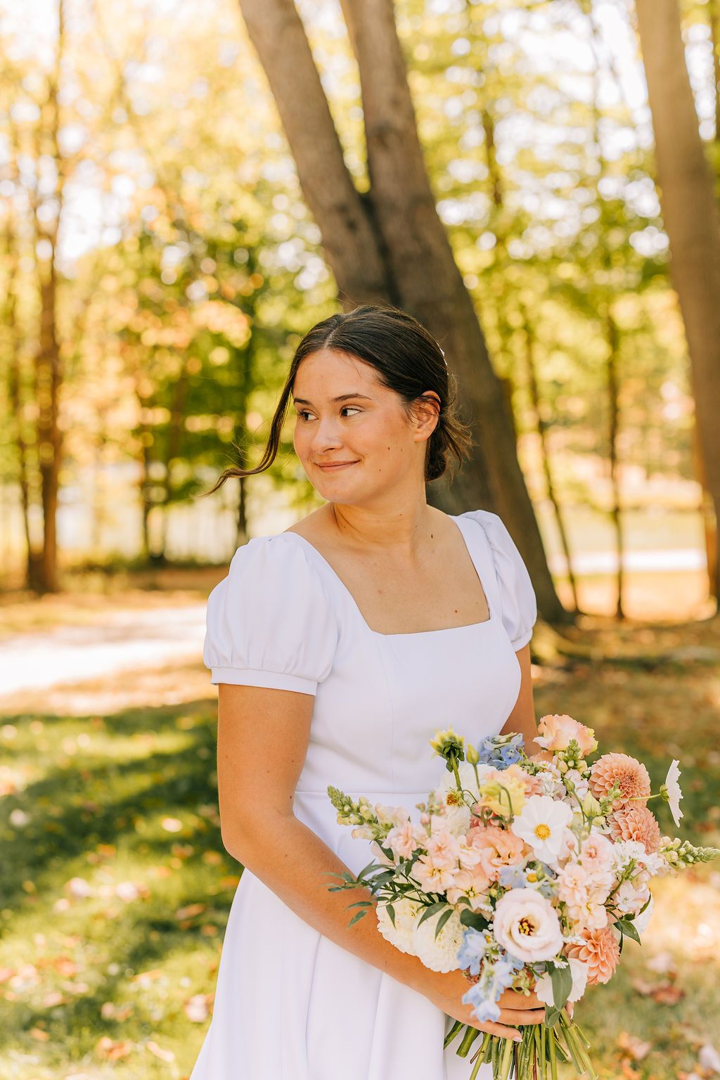 A person in a white dress holds a beautiful bouquet of pink and white flowers in a sunlit forest setting.