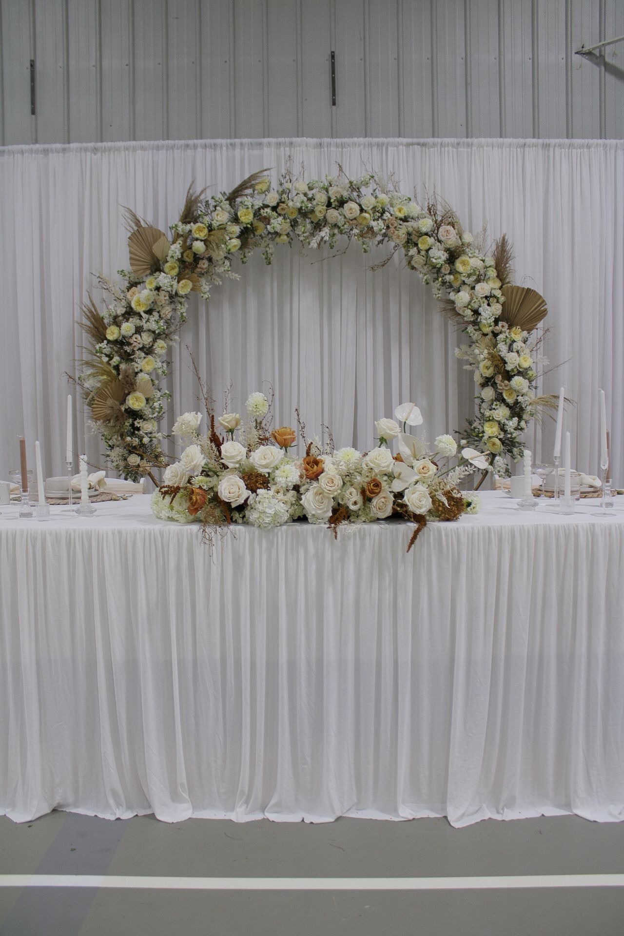 White floral wedding arch arrangements with roses and greenery adorn a reception head table against white draping.