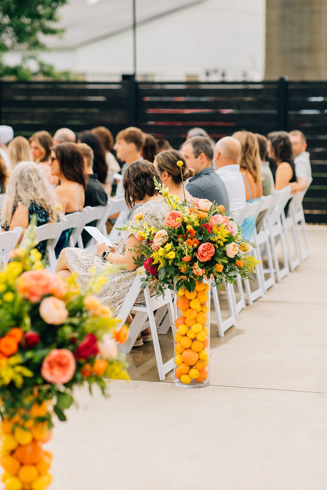 Wedding ceremony seating with white chairs and vibrant citrus floral arrangements featuring oranges and colorful blooms.
