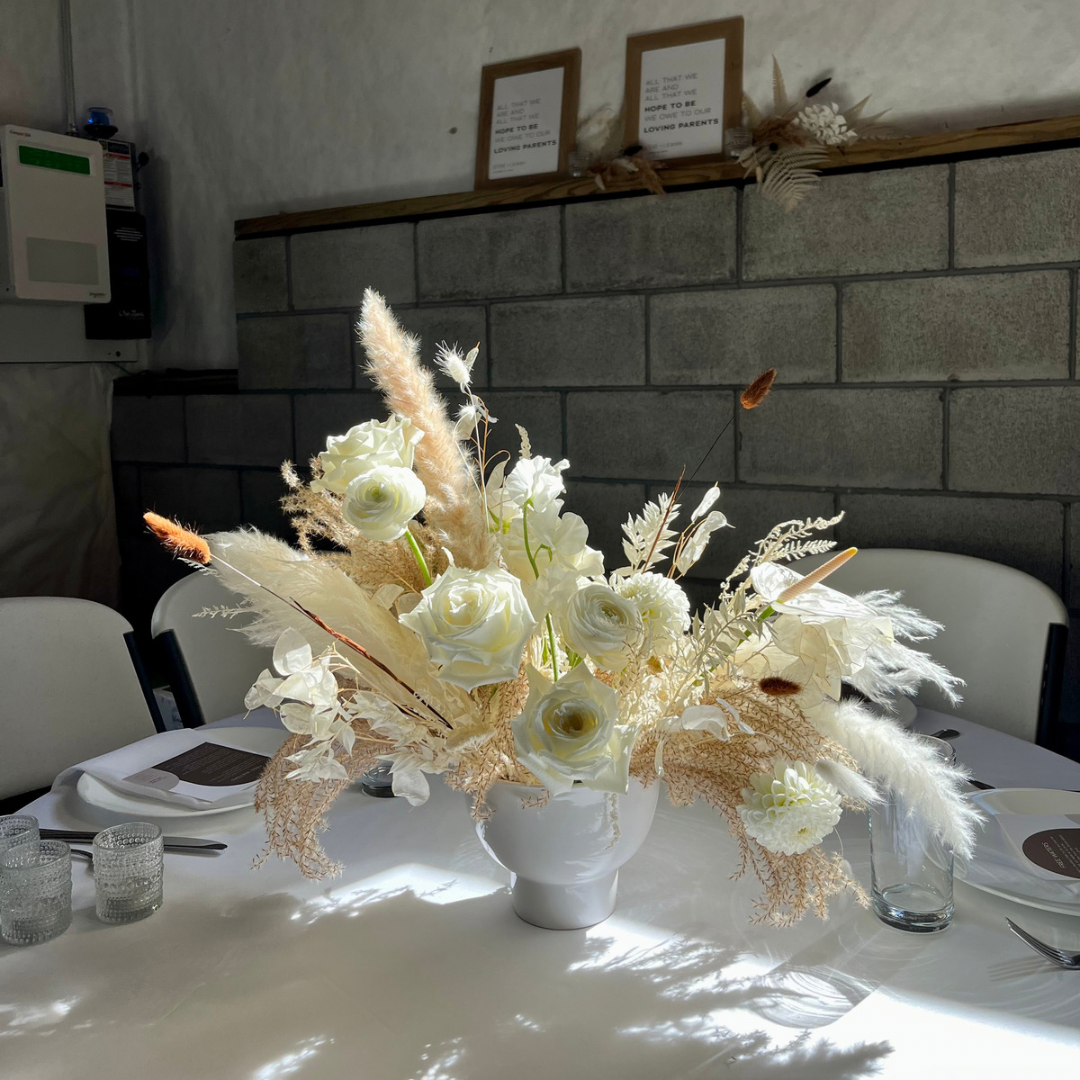 An elegant white floral centerpiece with feathers and dried elements in a white vase on a table setting.