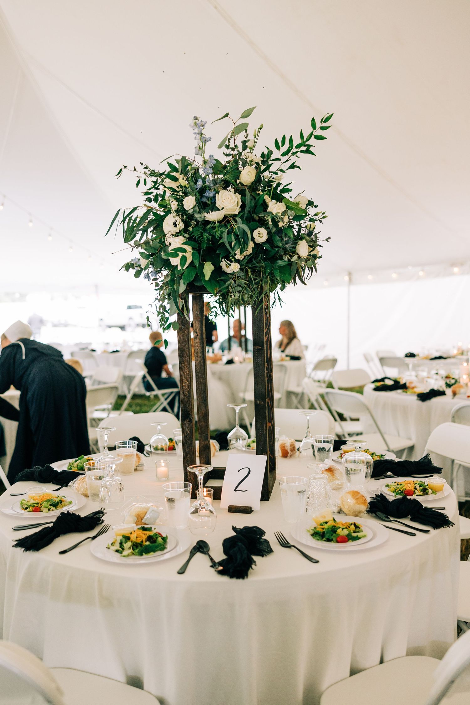 Round wedding table with tall floral centerpiece featuring white flowers and greenery under a white tent.