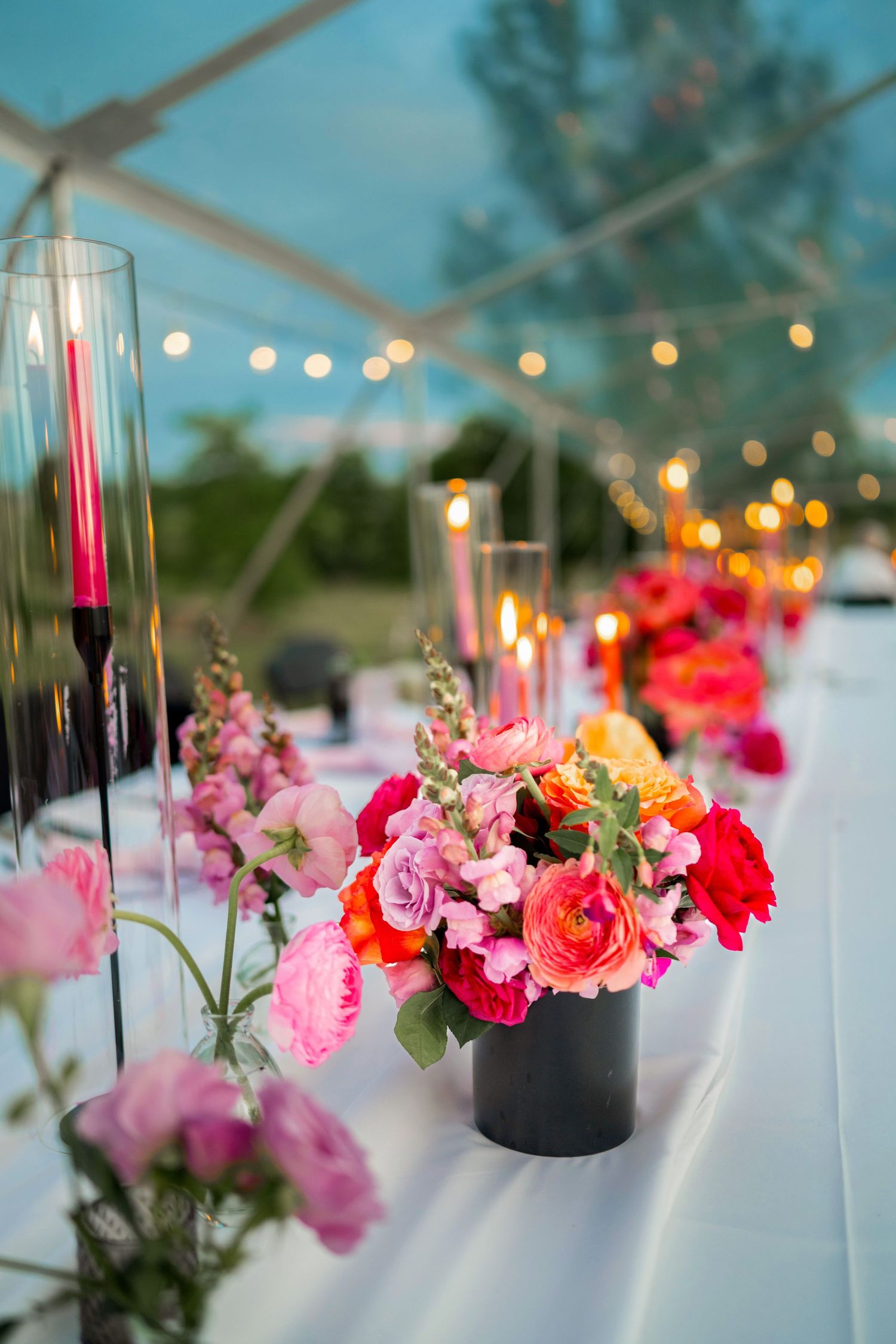 Vibrant pink and coral flower arrangements with glowing string lights at dusk under outdoor tent.