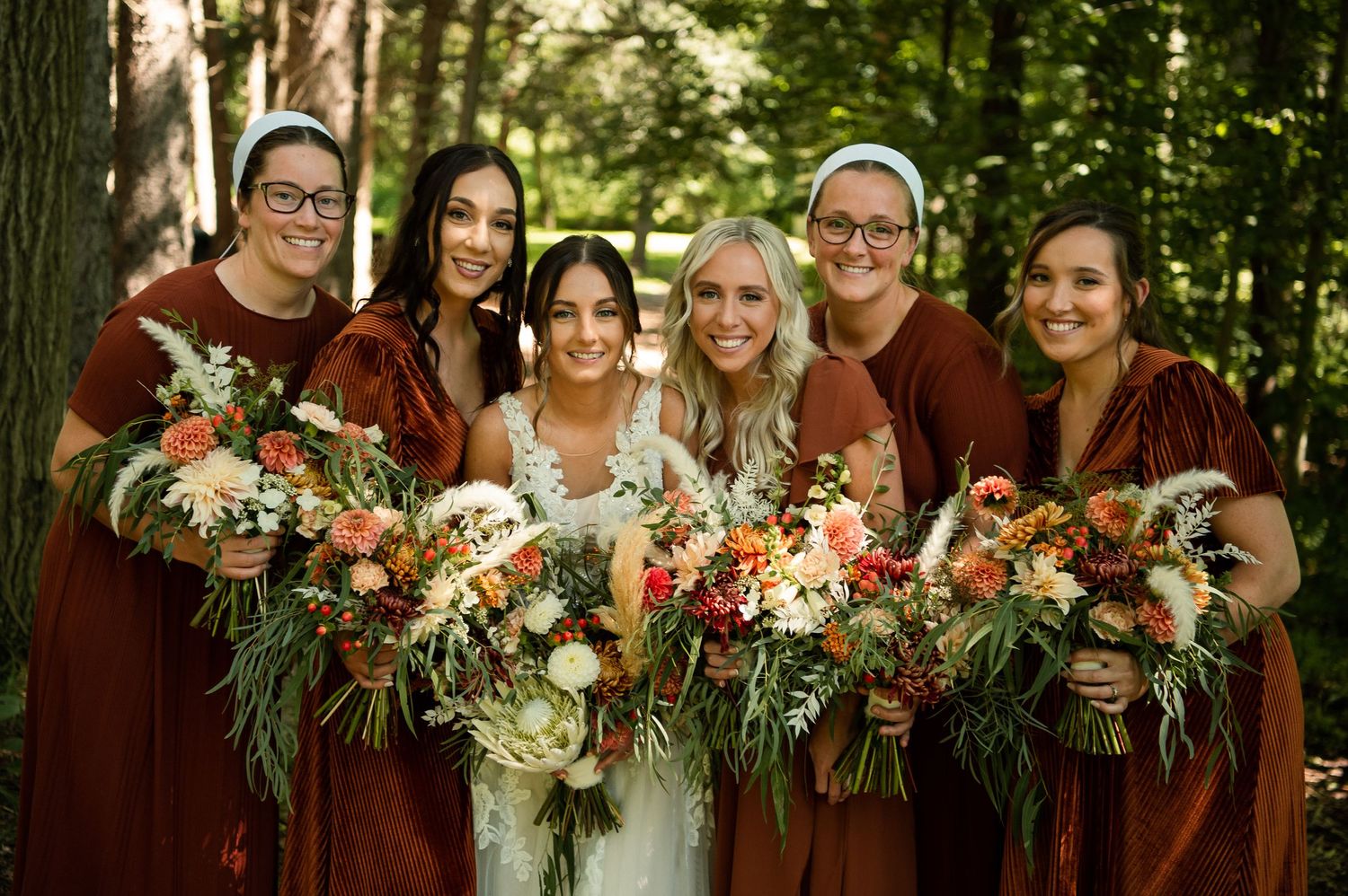 Bridal party in burgundy dresses poses with colorful autumn bouquets in forest setting.