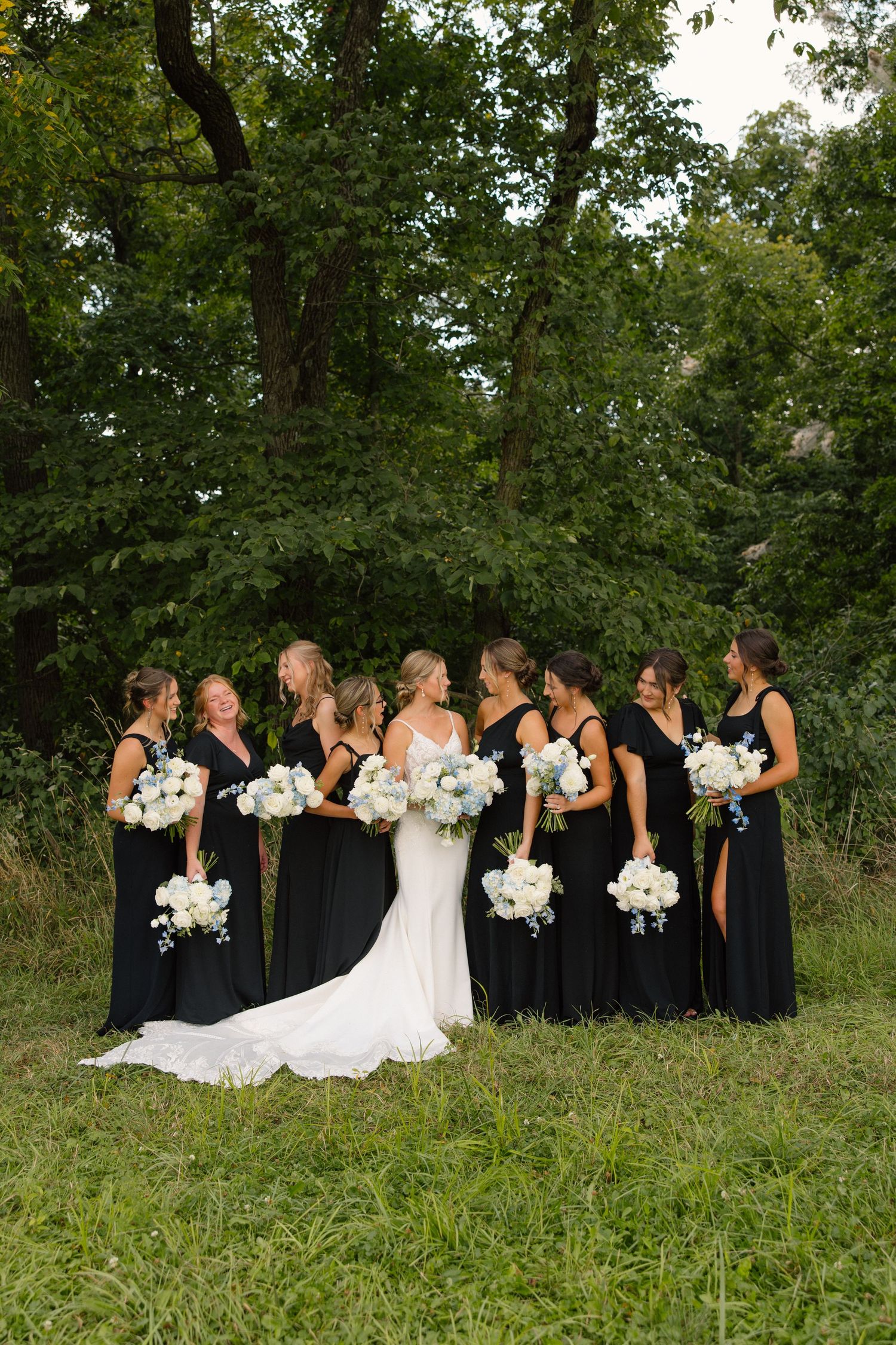 Bridal party in black dresses poses together outdoors holding white floral bouquets.