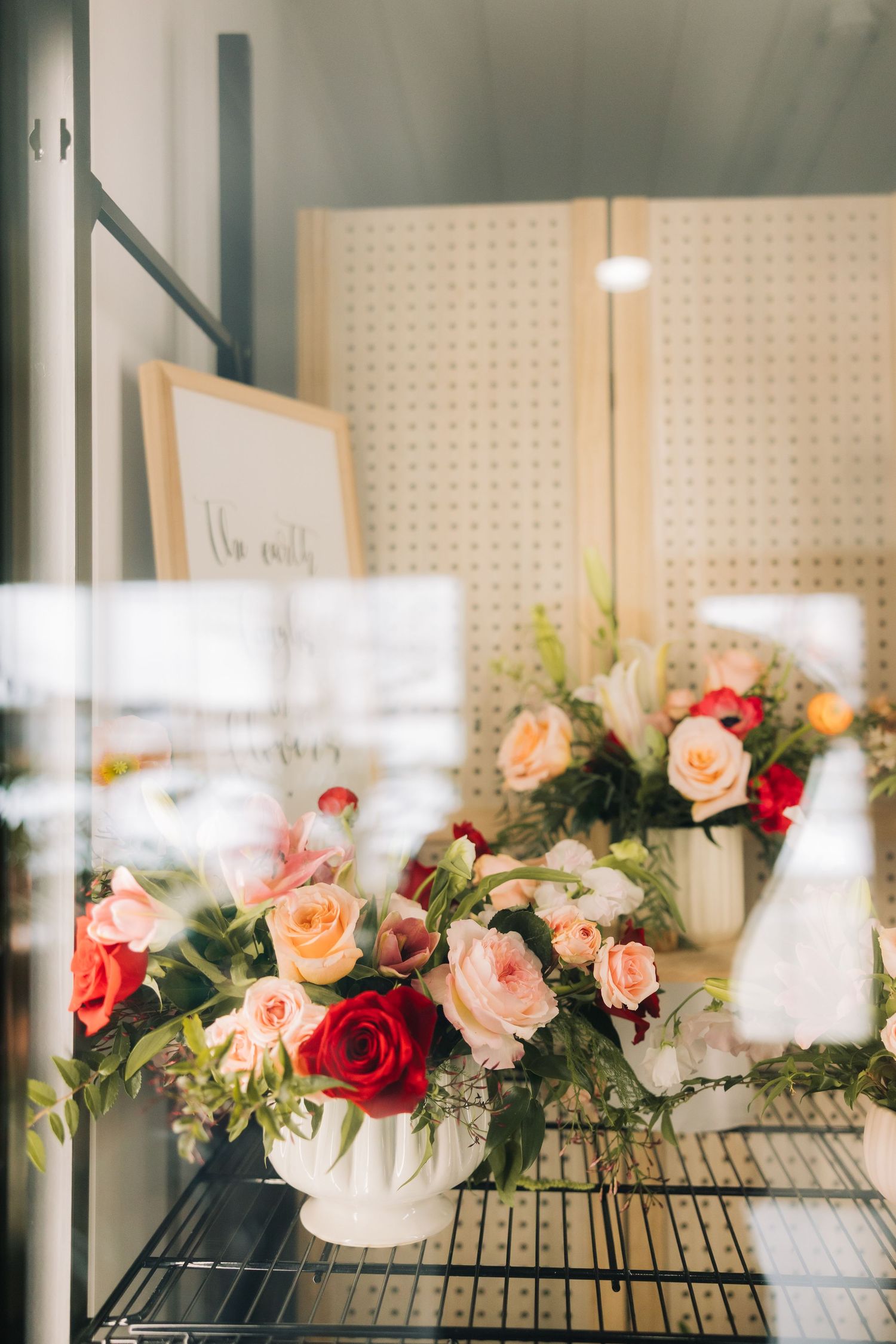 Beautiful floral arrangement with pink and red roses displayed in white vases on a shelf with pegboard backdrop.